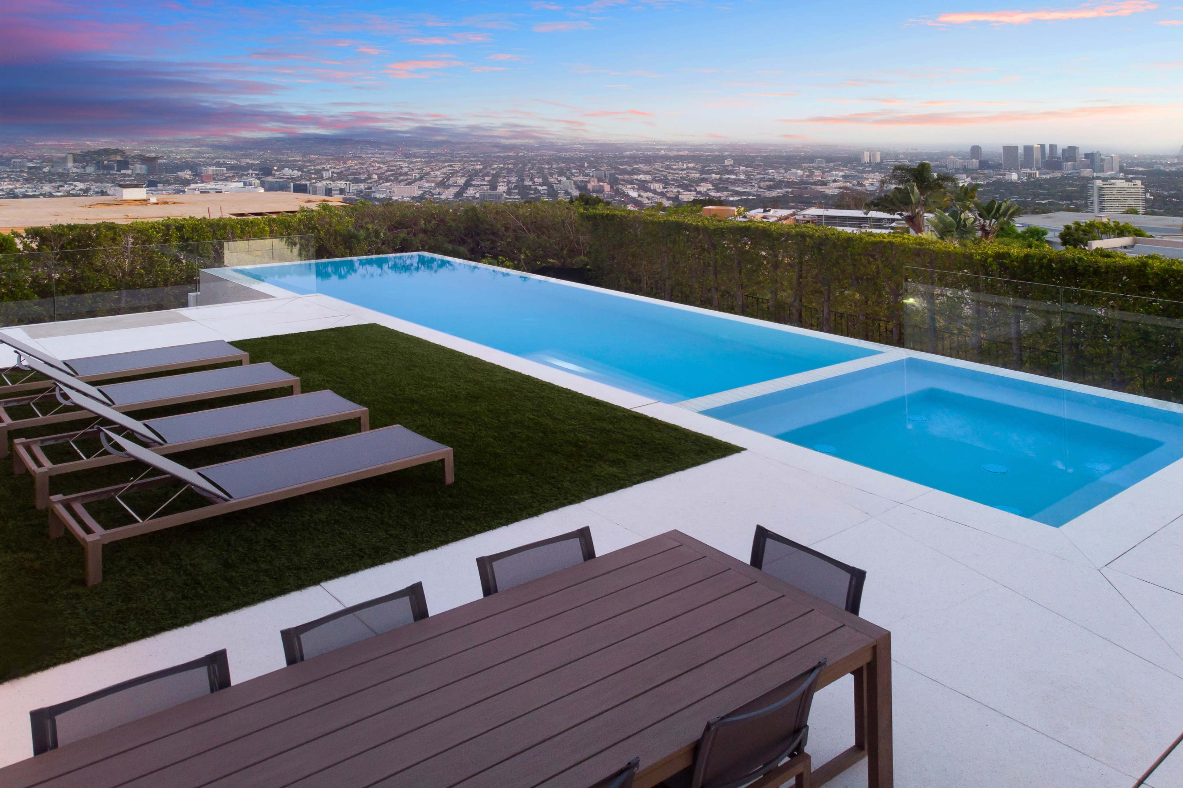 A modern outdoor space features a swimming pool, lounge chairs, and a dining table, overlooking a city skyline at sunset.