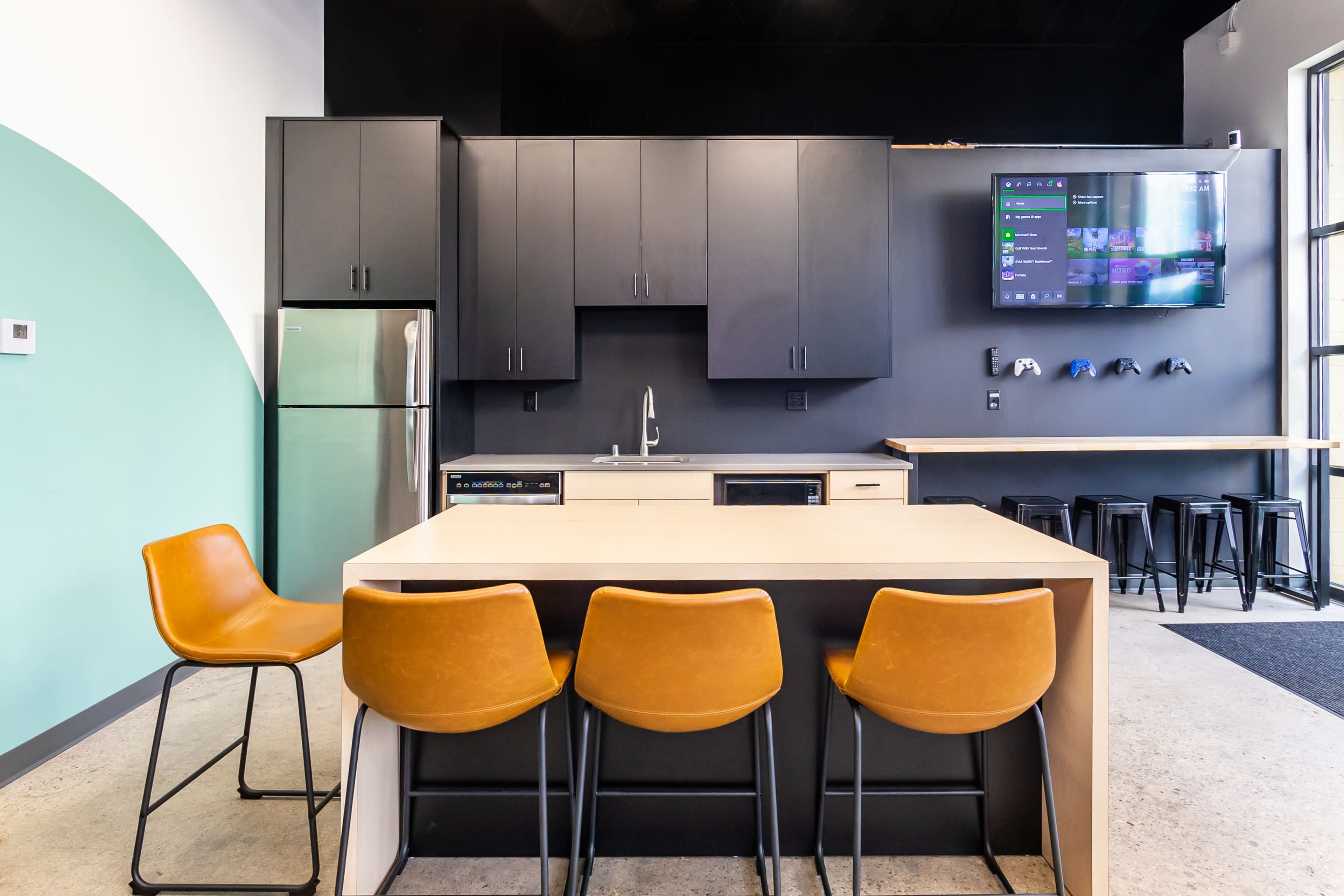 A modern kitchen area with a central island, brown bar stools, black cabinetry, and a television mounted on the wall.