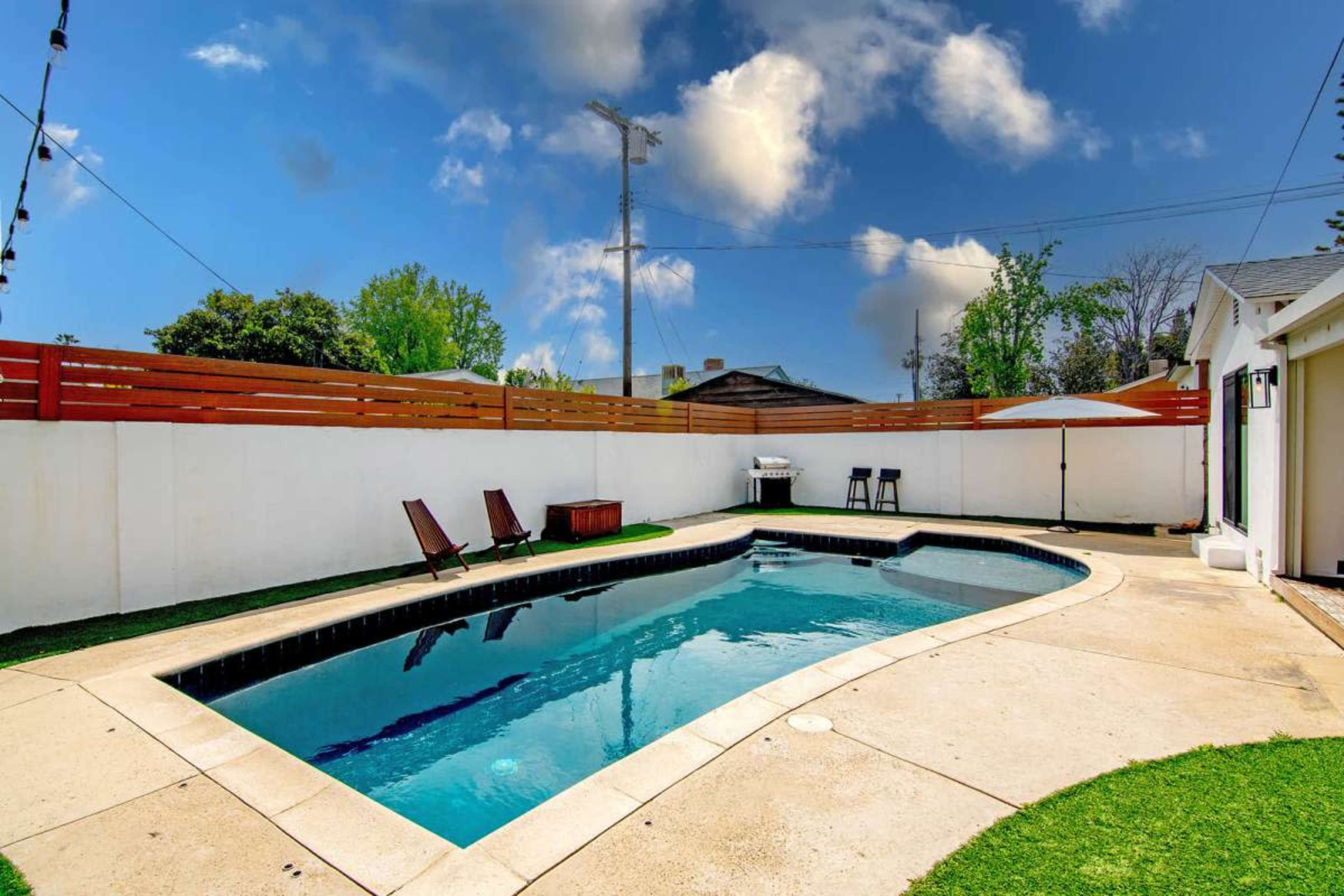 The image shows a rectangular swimming pool surrounded by a patio area with lounge chairs and a small table, enclosed by a wooden fence and a grassy border.