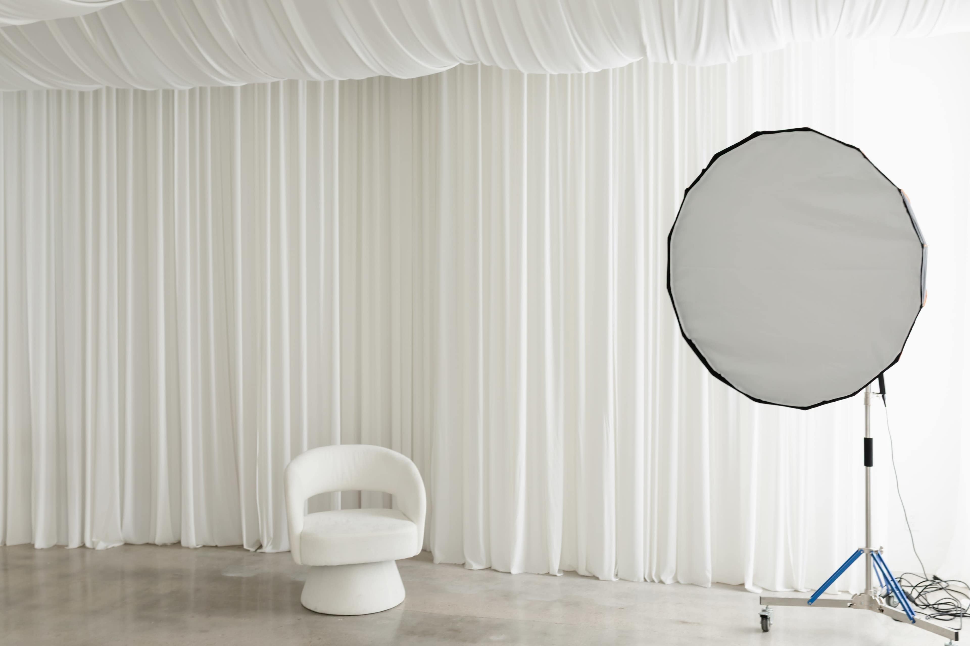 A white, minimalist chair sits against a backdrop of white drapery, with a large circular reflector positioned nearby.