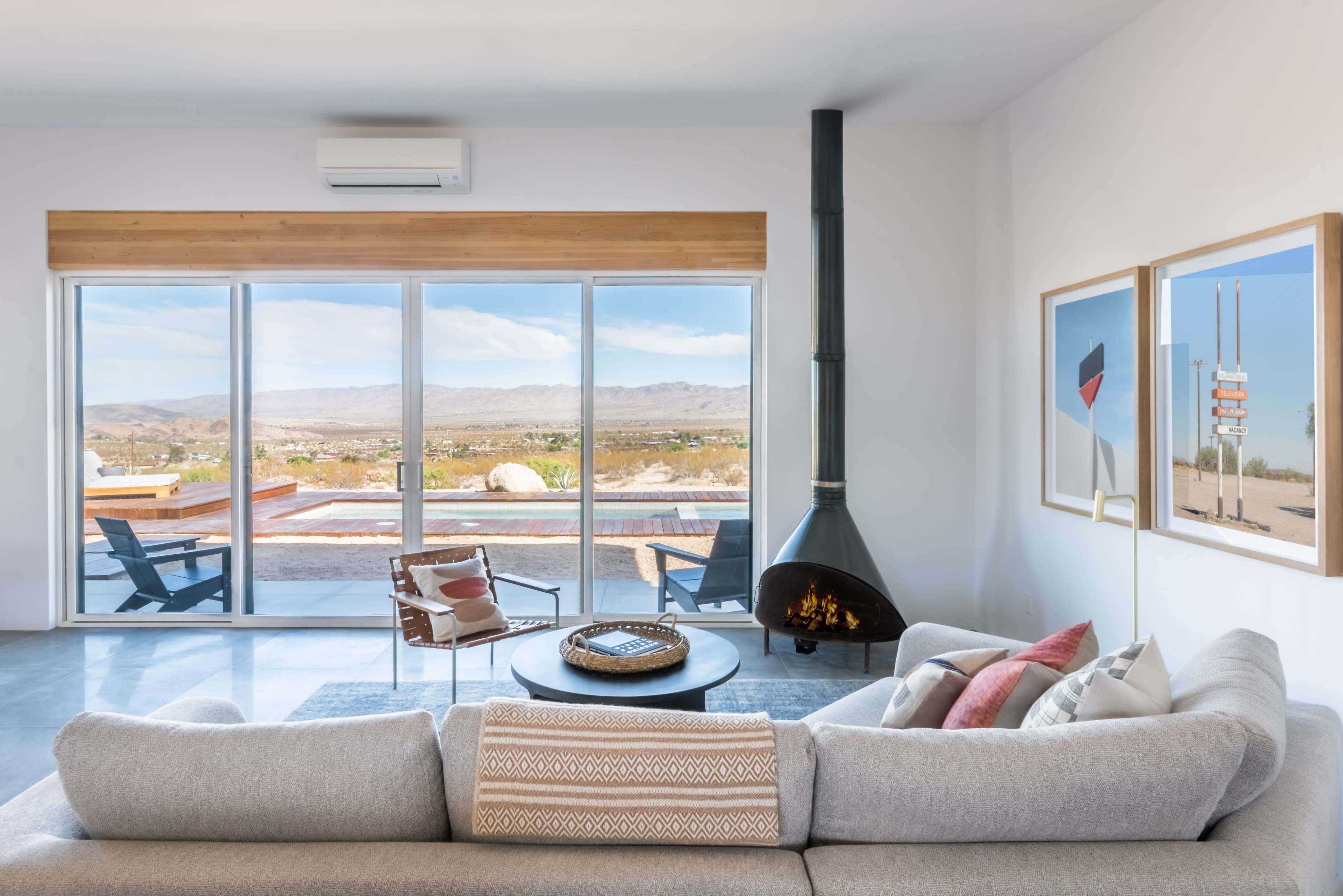 The image shows a modern, open-concept living room featuring a large window with a panoramic view of a desert landscape, a cozy sofa, a round coffee table, and a sleek black fireplace.