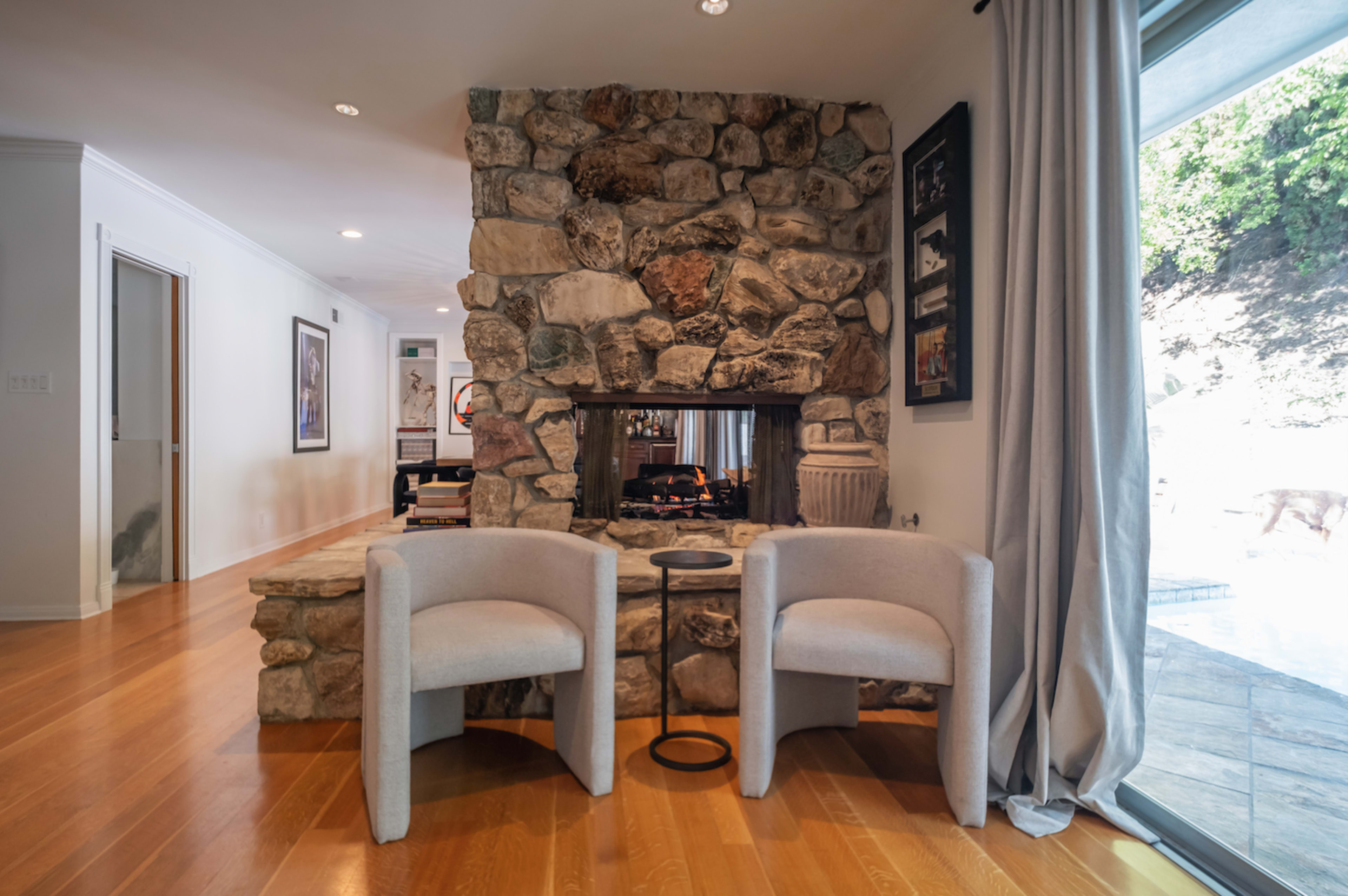A living room with a stone fireplace, two light-colored chairs, and large glass doors leading to an outdoor area.