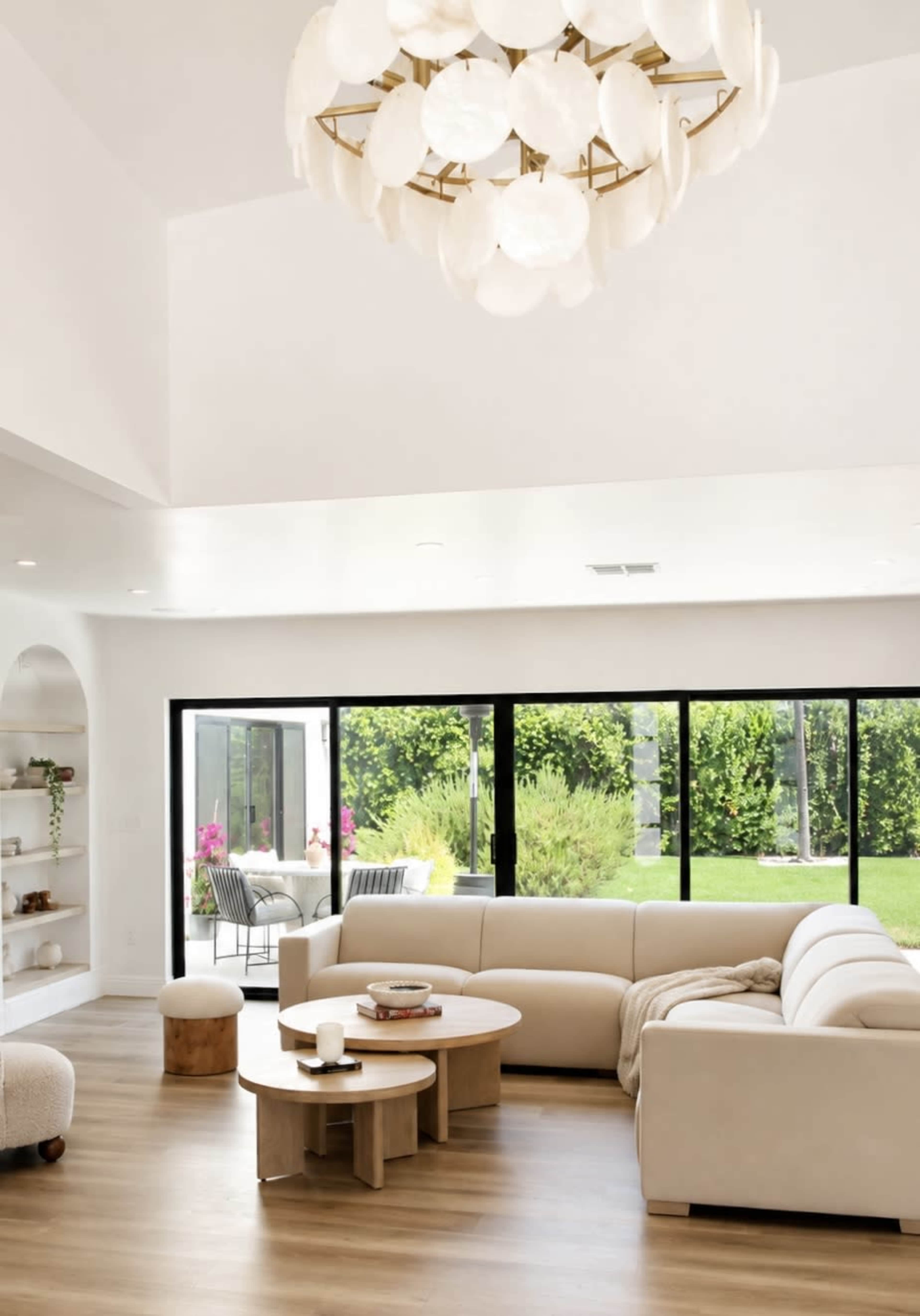 The image shows a modern living room with a large light fixture, a beige sectional sofa, and large windows overlooking a green garden.