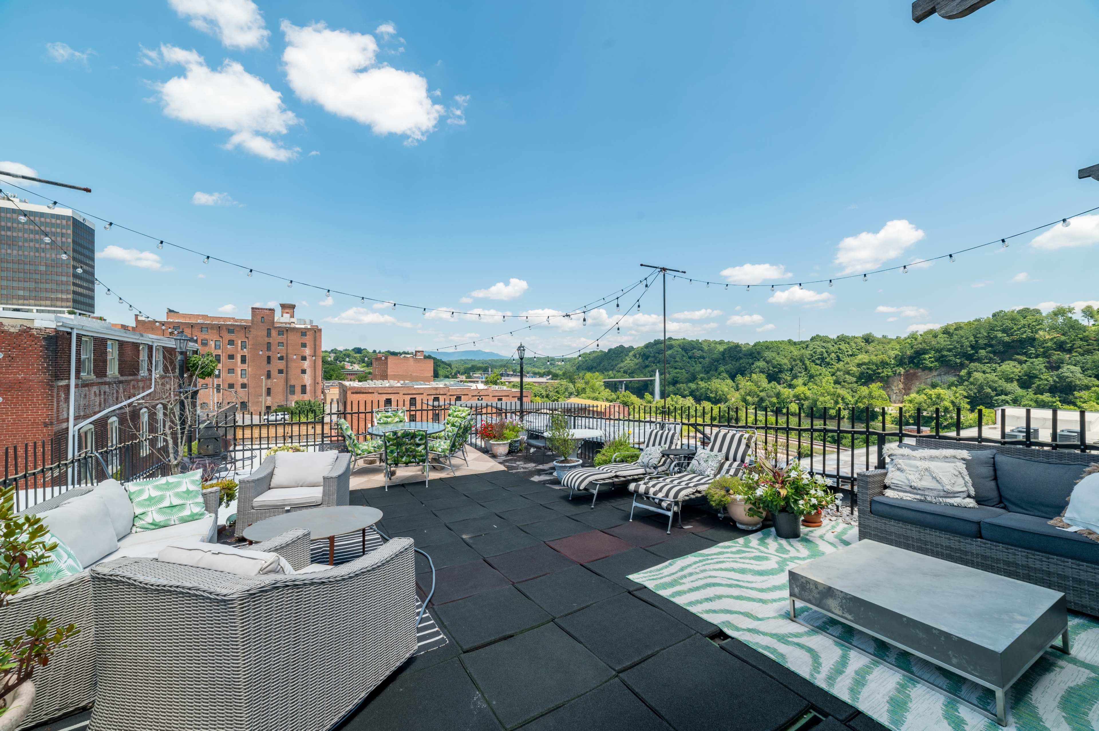 A spacious rooftop terrace features seating areas, decorative plants, and a view of distant hills under a clear sky with scattered clouds.