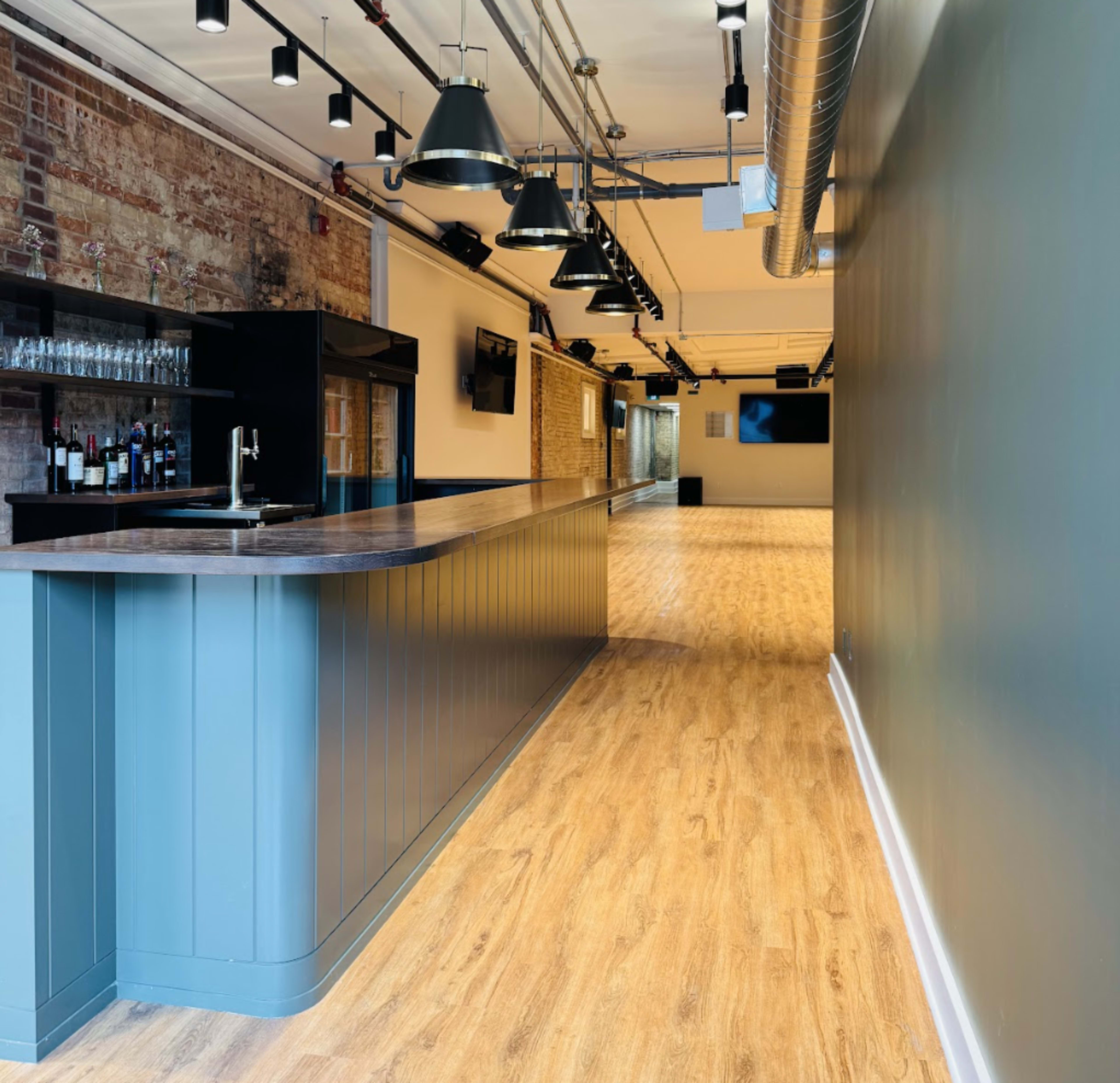 The image shows a long, narrow bar area with a wooden counter, light hardwood flooring, and exposed brick walls, leading into a spacious, open room with track lighting and wall-mounted screens.