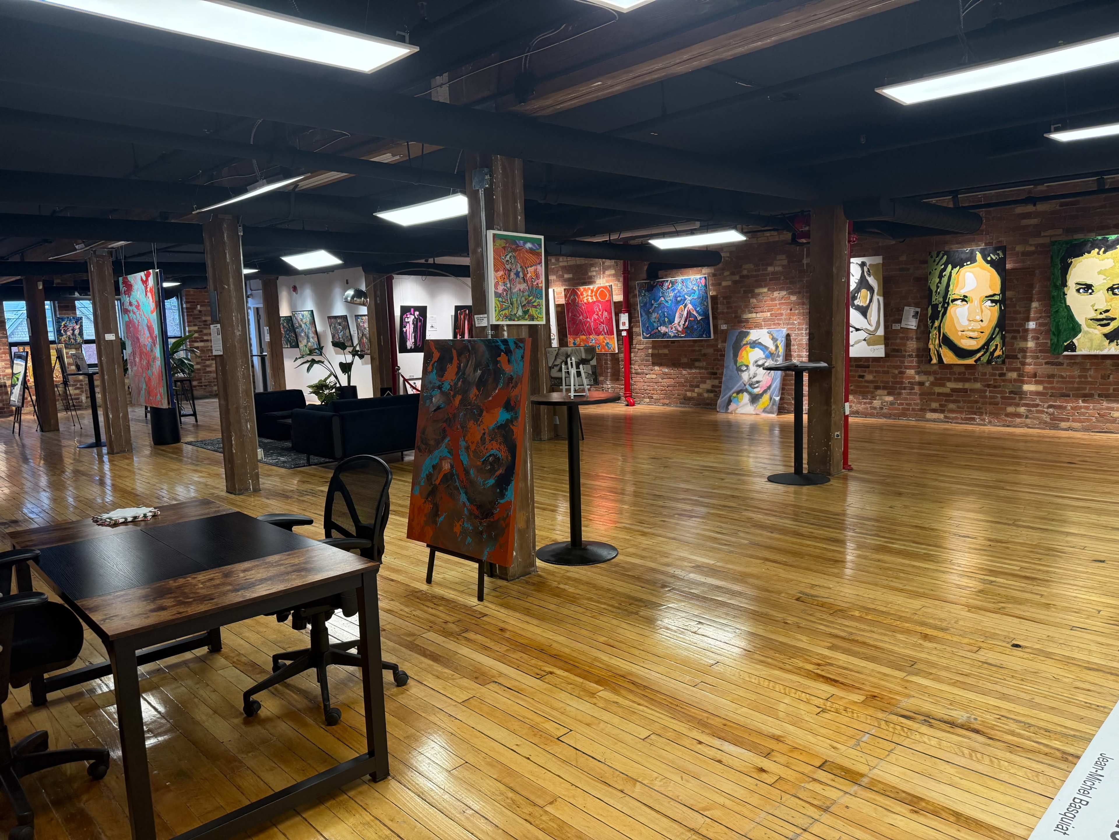 The image shows an art gallery featuring colorful paintings displayed on brick walls, with wooden floors and modern furniture.