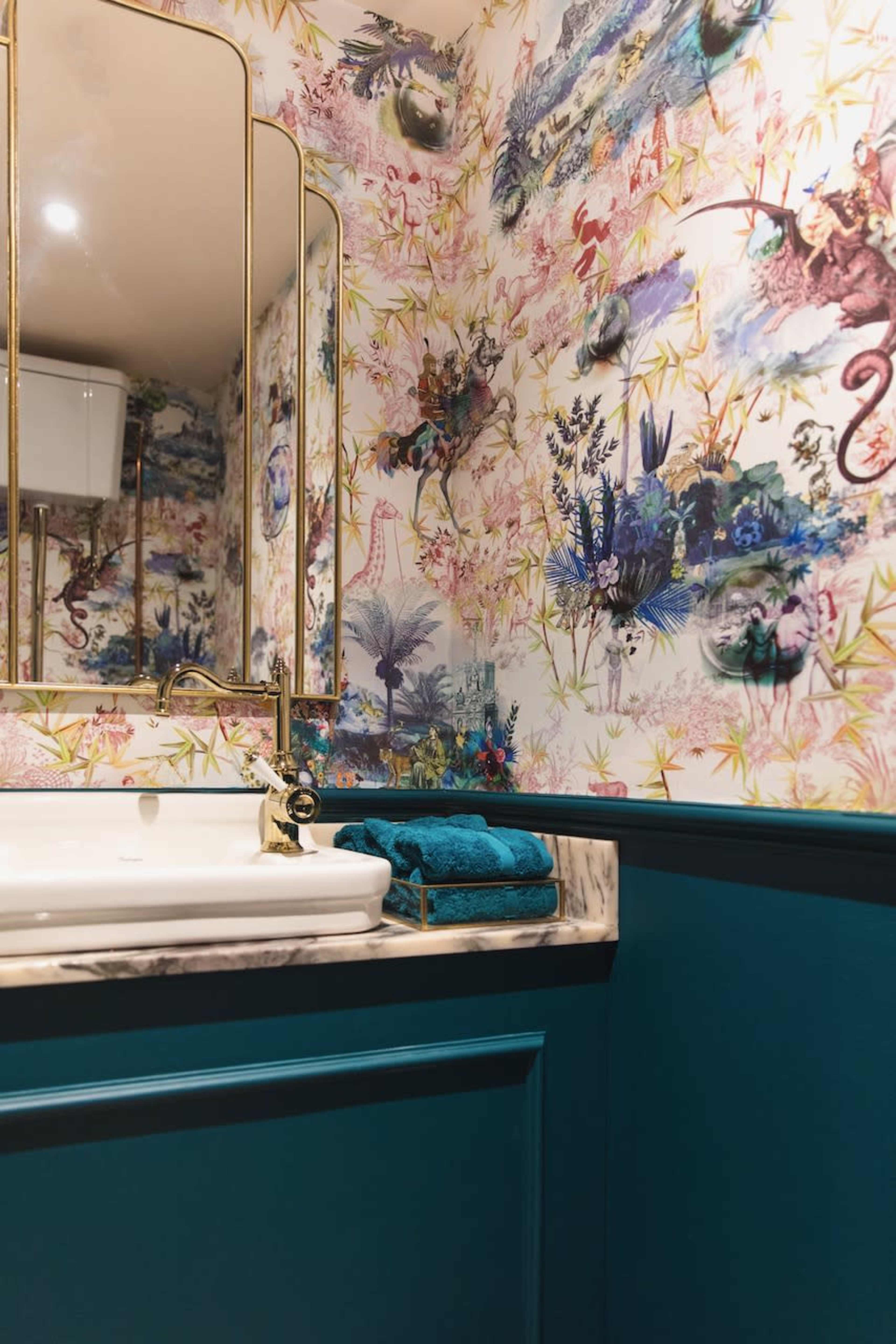 A bathroom with a marble sink and a patterned wallpaper featuring colorful floral and animal designs, complemented by a teal-painted lower wall and neatly stacked blue towels.