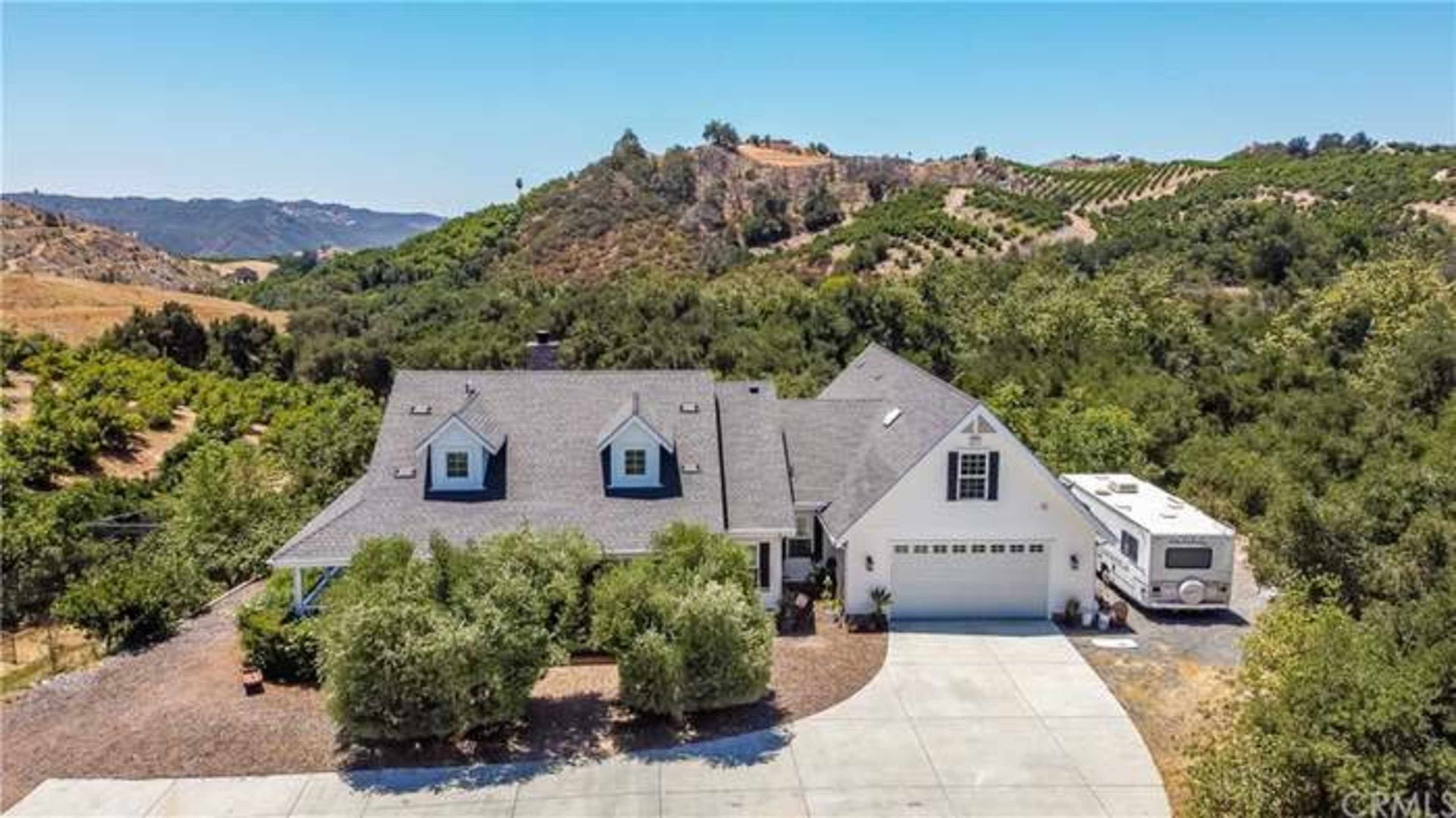 A large house with a gray roof, situated on a paved driveway, surrounded by trees and hills in the background.