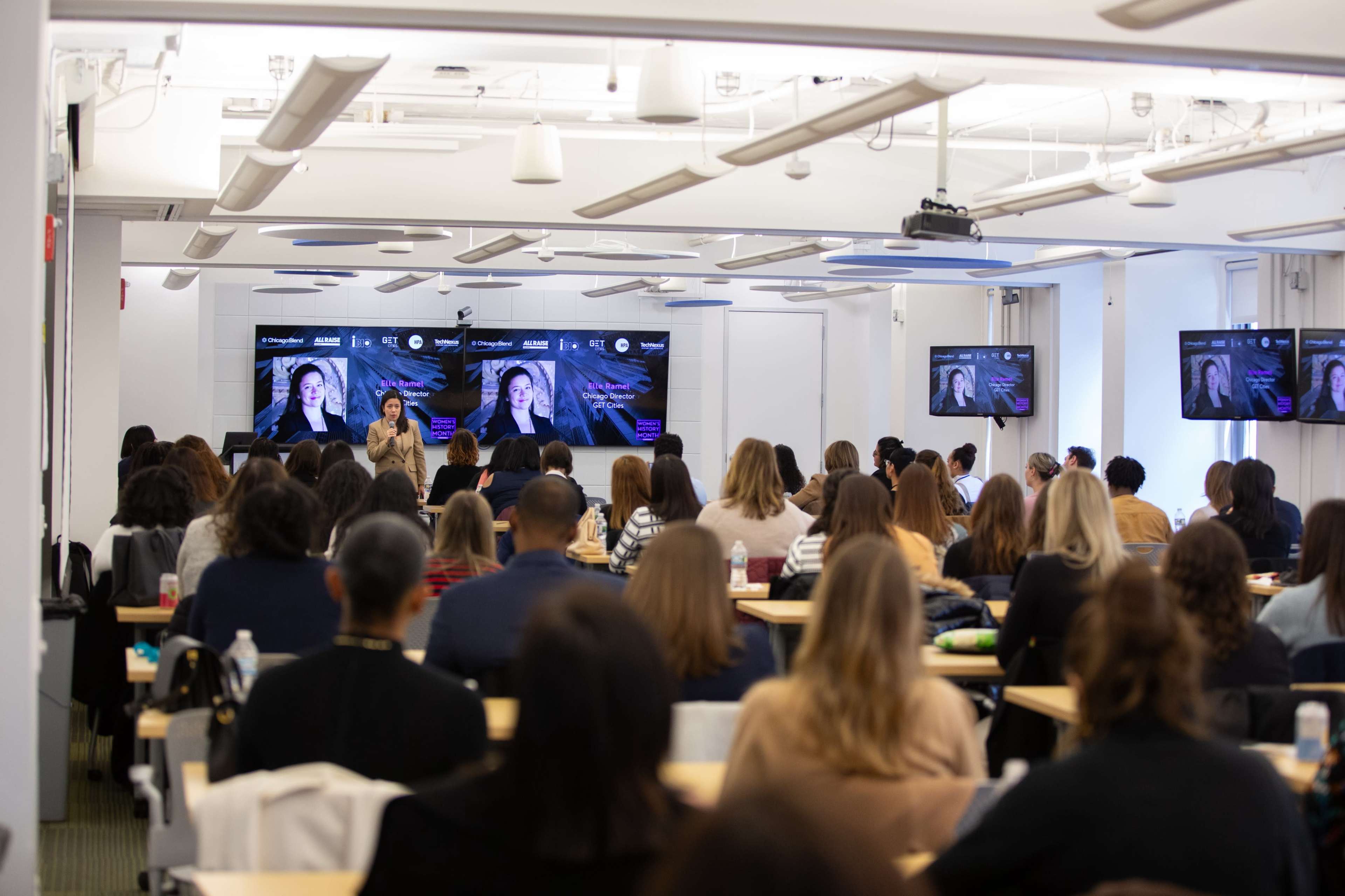 A group of people sits in a classroom setting, facing a presenter and screens displaying visuals.
