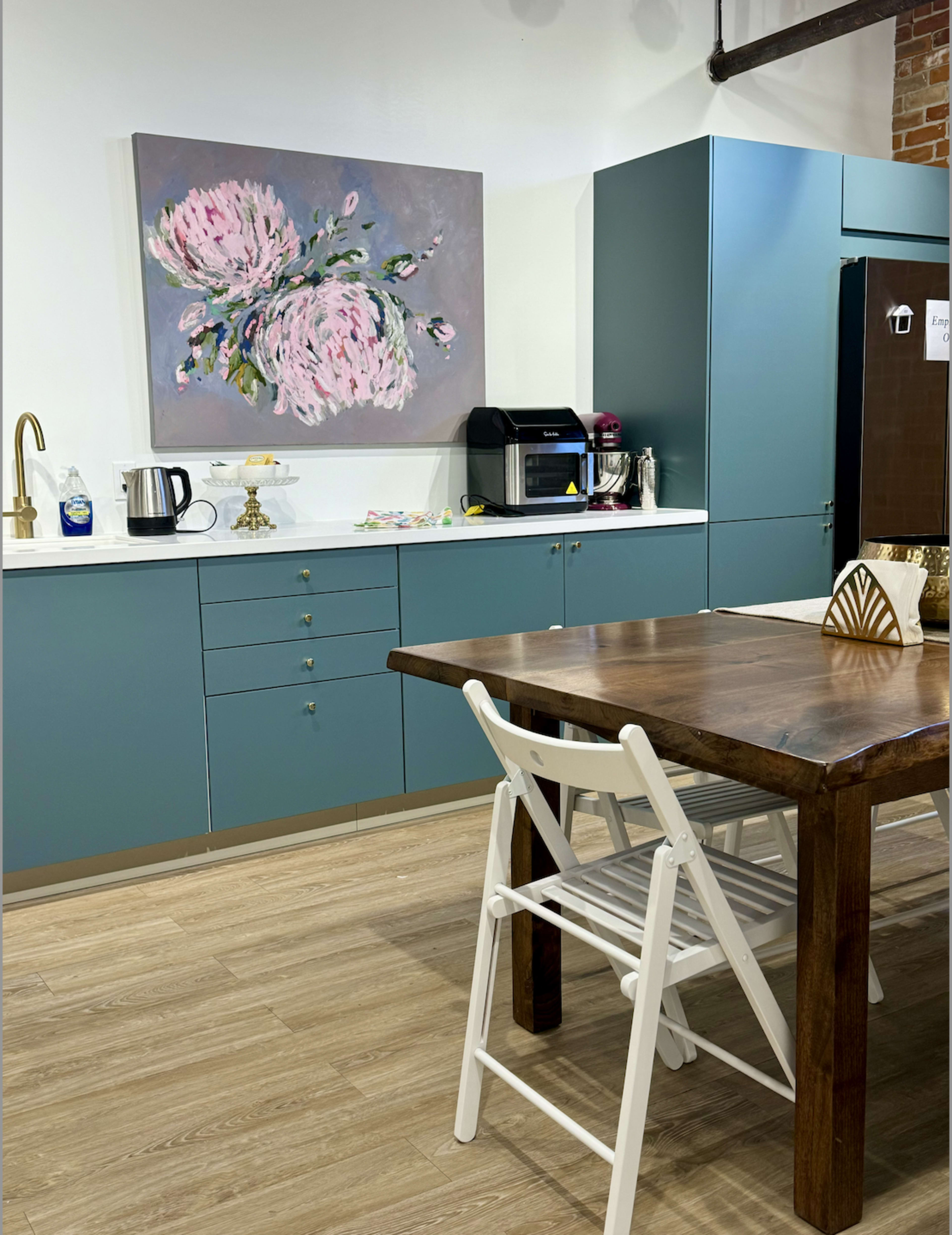 The image shows a modern kitchen with teal cabinets, a wooden dining table, a coffee machine, and a large floral painting on the wall.