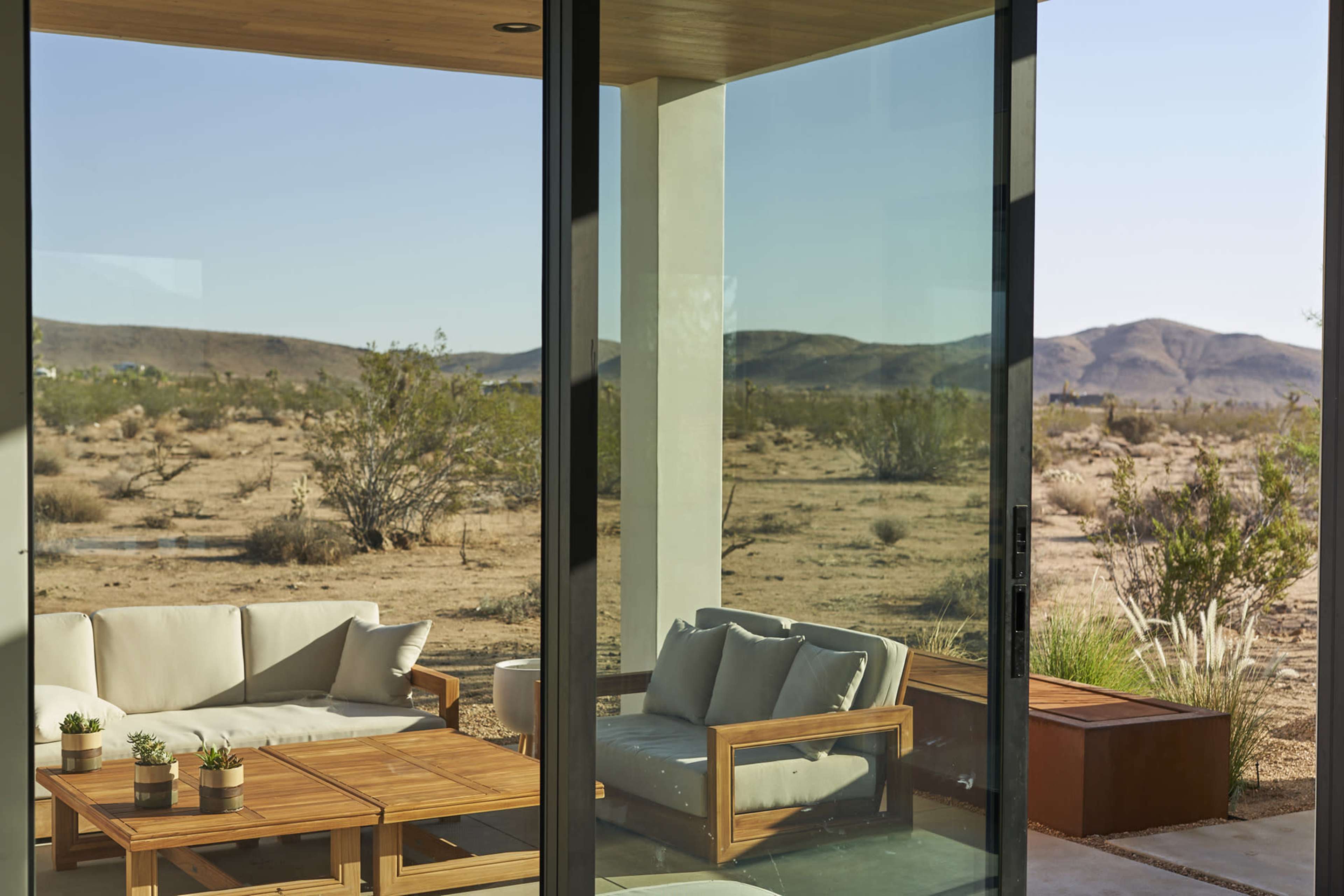 A modern outdoor seating area with wooden furniture and potted plants overlooks a desert landscape through large glass windows.
