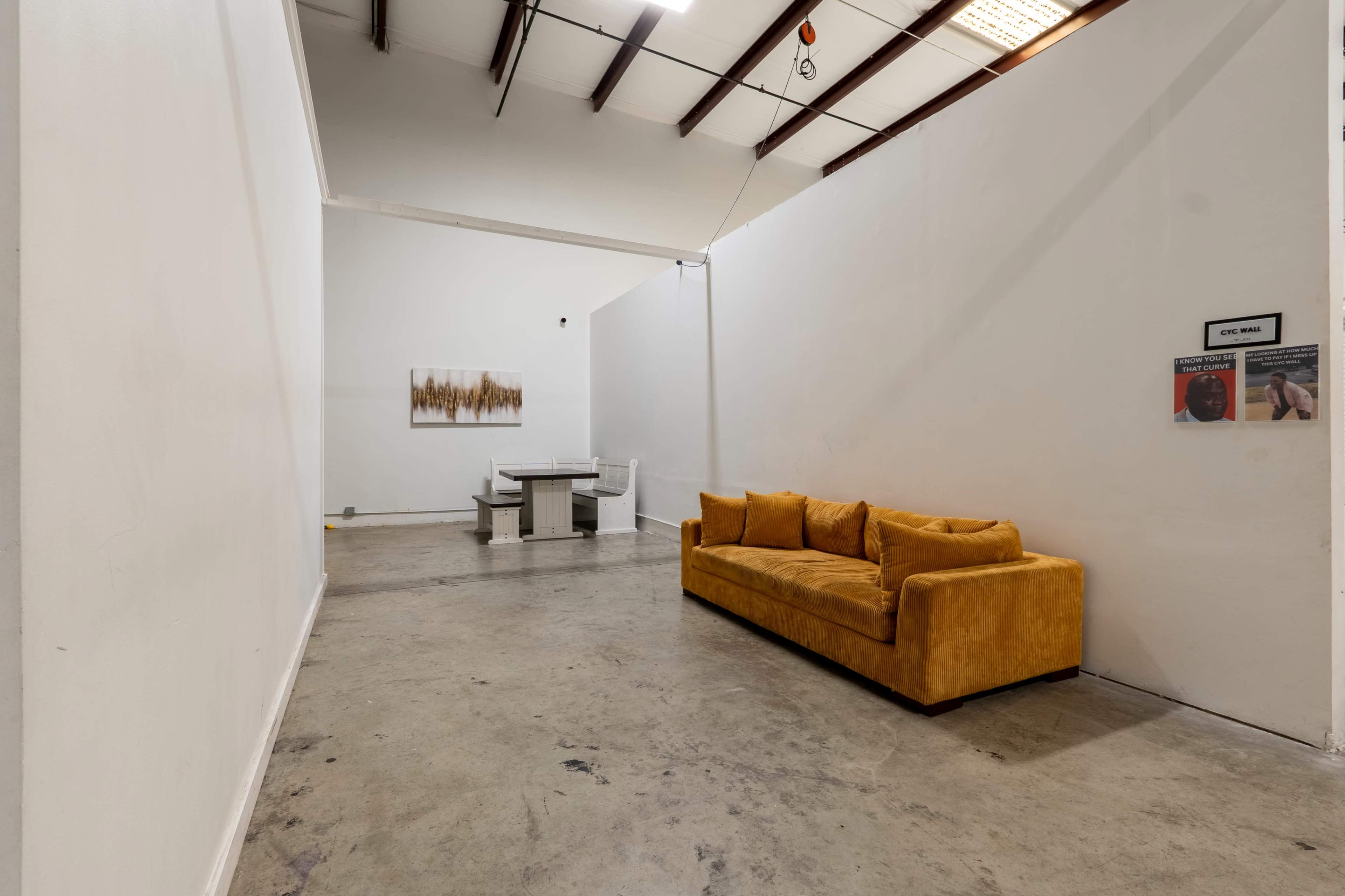 The image shows a spacious, minimal interior featuring a yellow sofa and a table with chairs in a well-lit room with white walls.