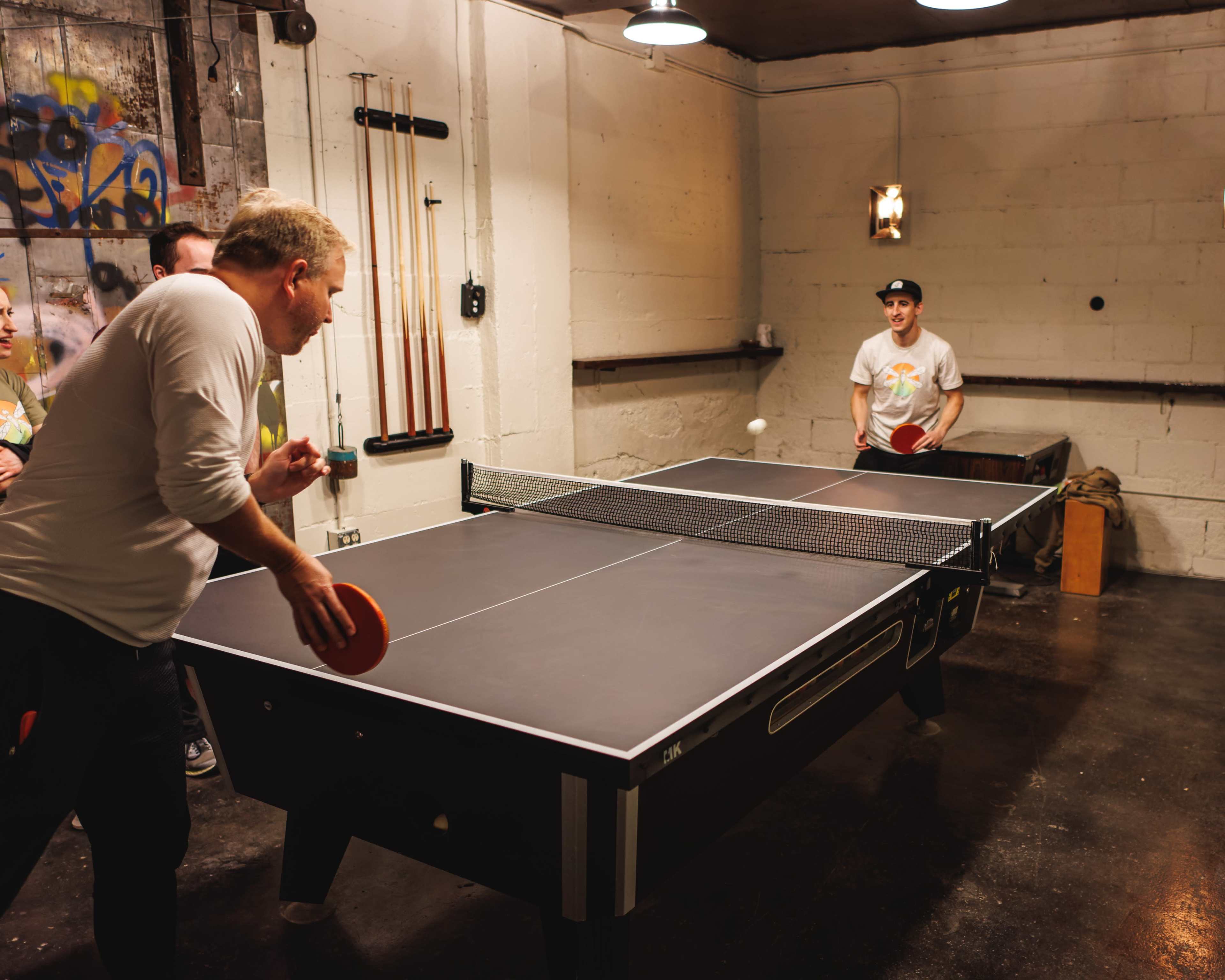 Two men play table tennis in a dimly lit indoor setting with exposed brick walls.