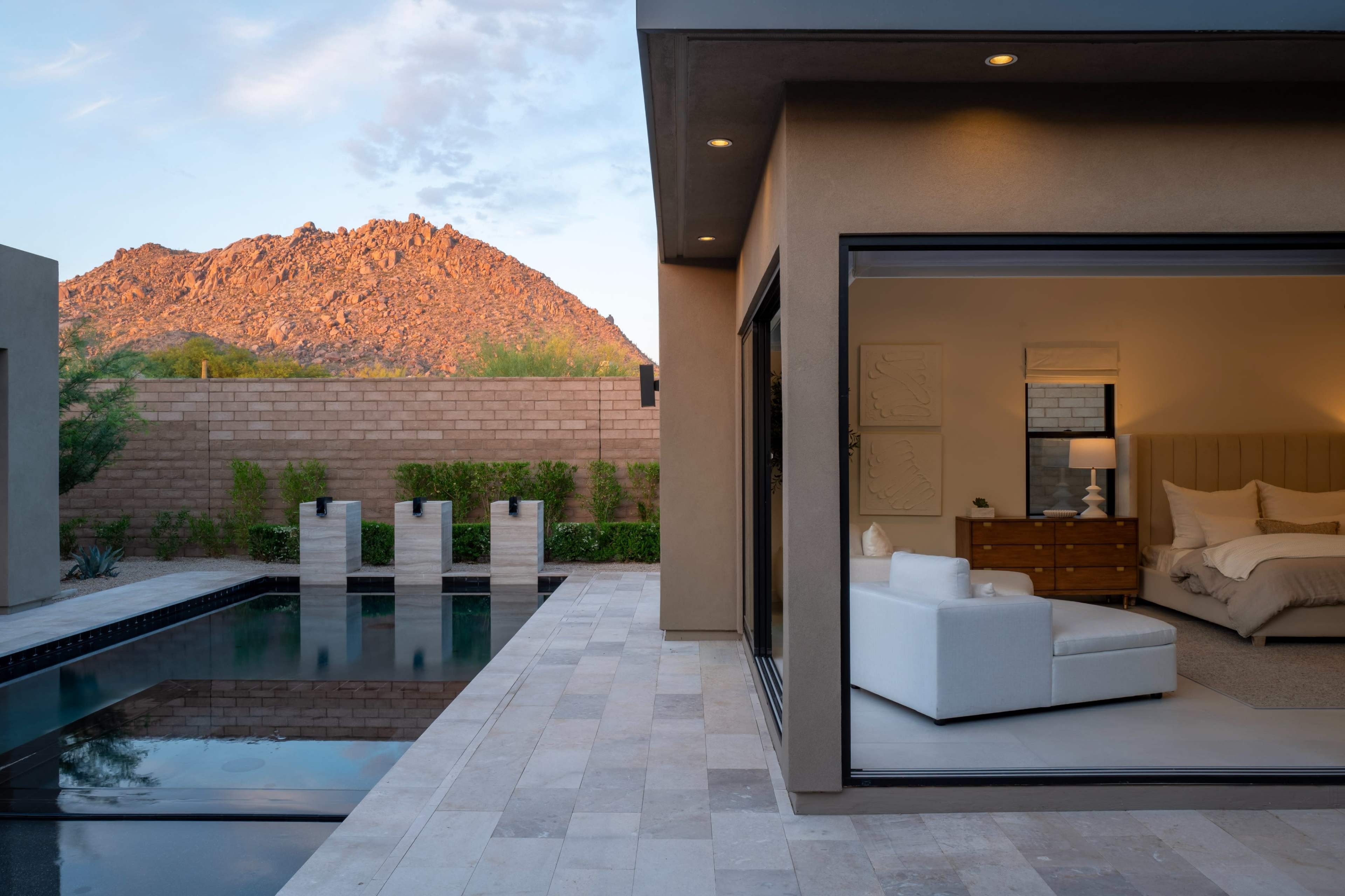 The image shows a modern home with sliding glass doors revealing a bedroom, a small swimming pool, and a mountain landscape in the background under a colorful sky.