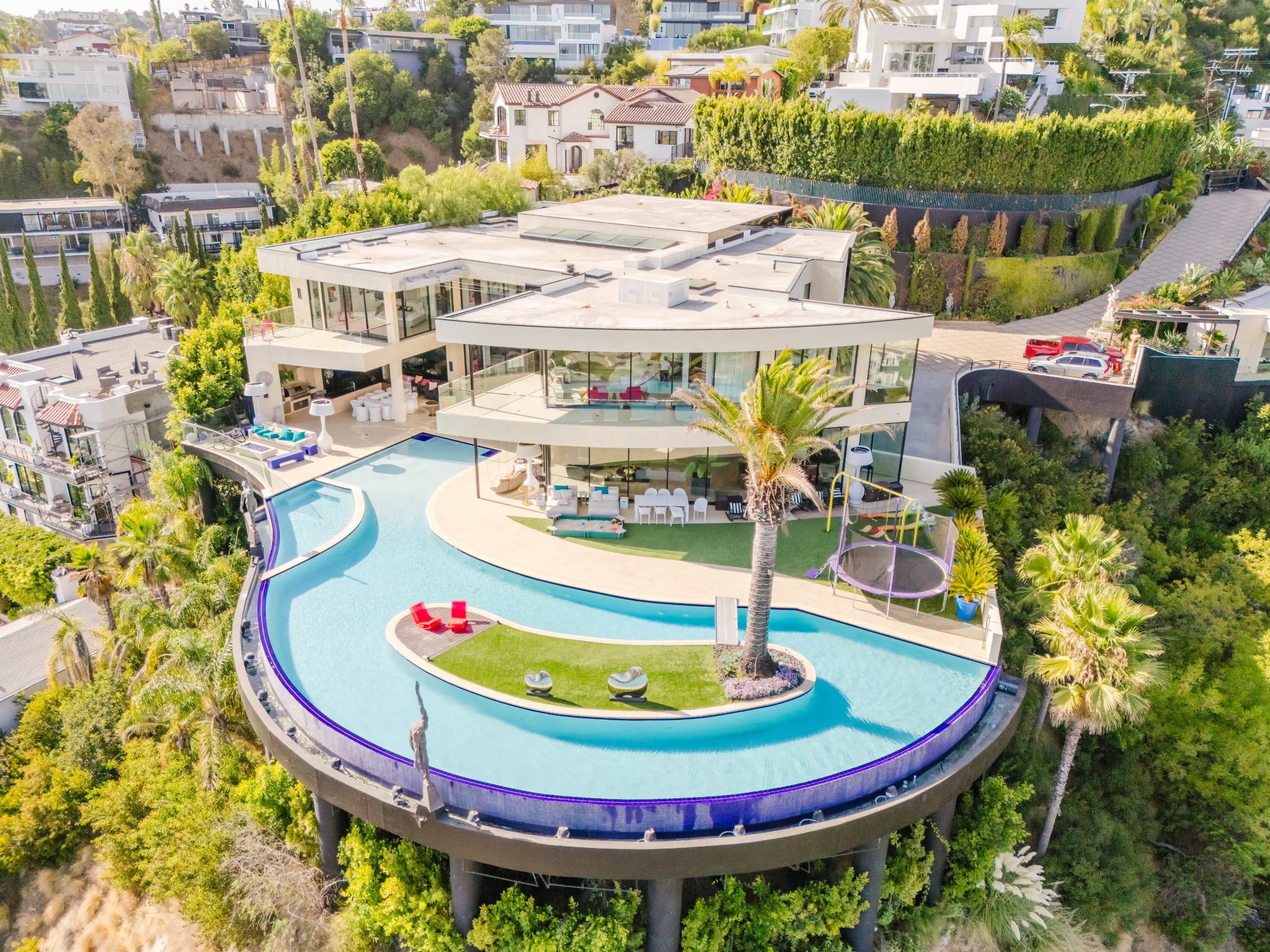 The image shows a modern, multi-level house with large glass windows, a circular swimming pool, and landscaped greenery surrounding it, situated on a hillside.