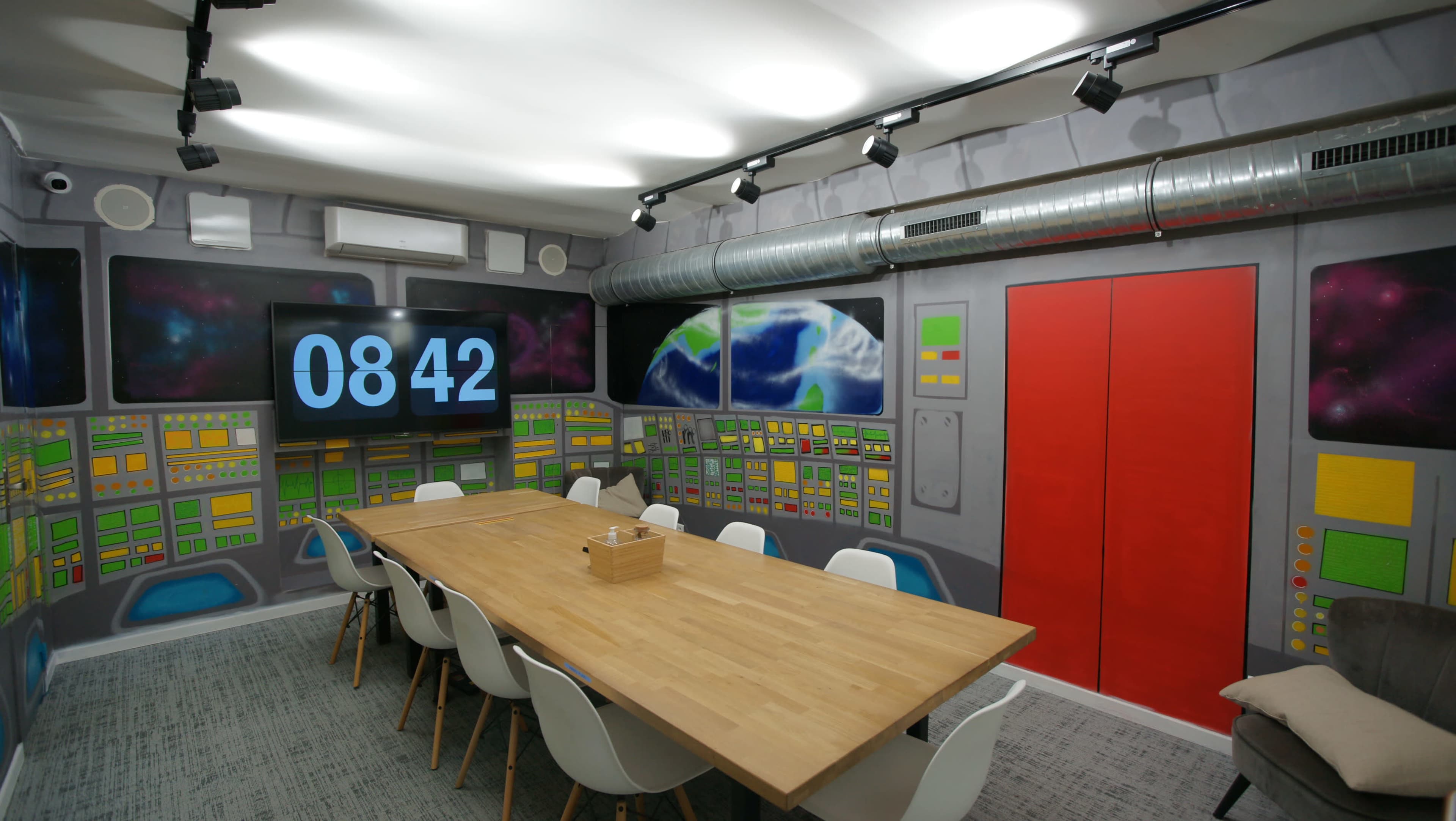 A modern conference room with a large wooden table, white chairs, and a wall decorated with a space-themed mural featuring control panels and a large digital clock displaying 08:42.