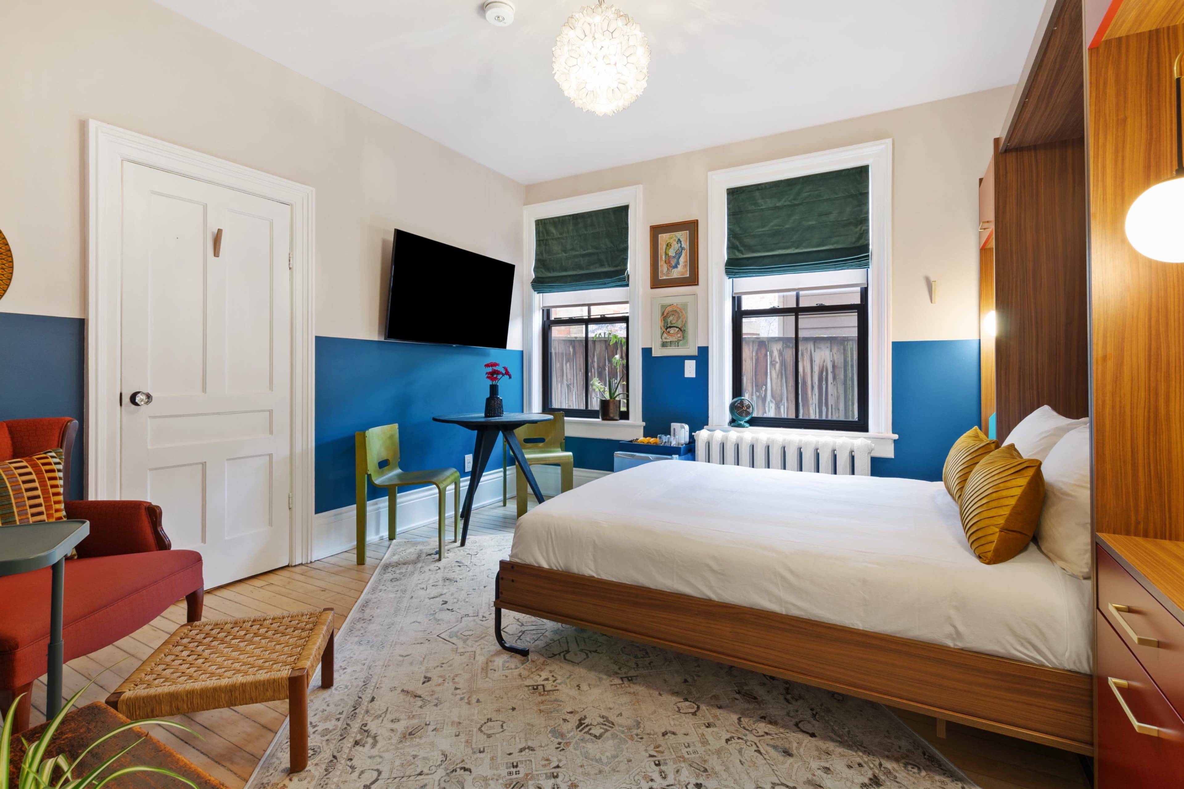 A modern bedroom features a bed, small table and chairs, a dresser, and a television mounted on the wall, with a mix of blue and beige paint on the walls.