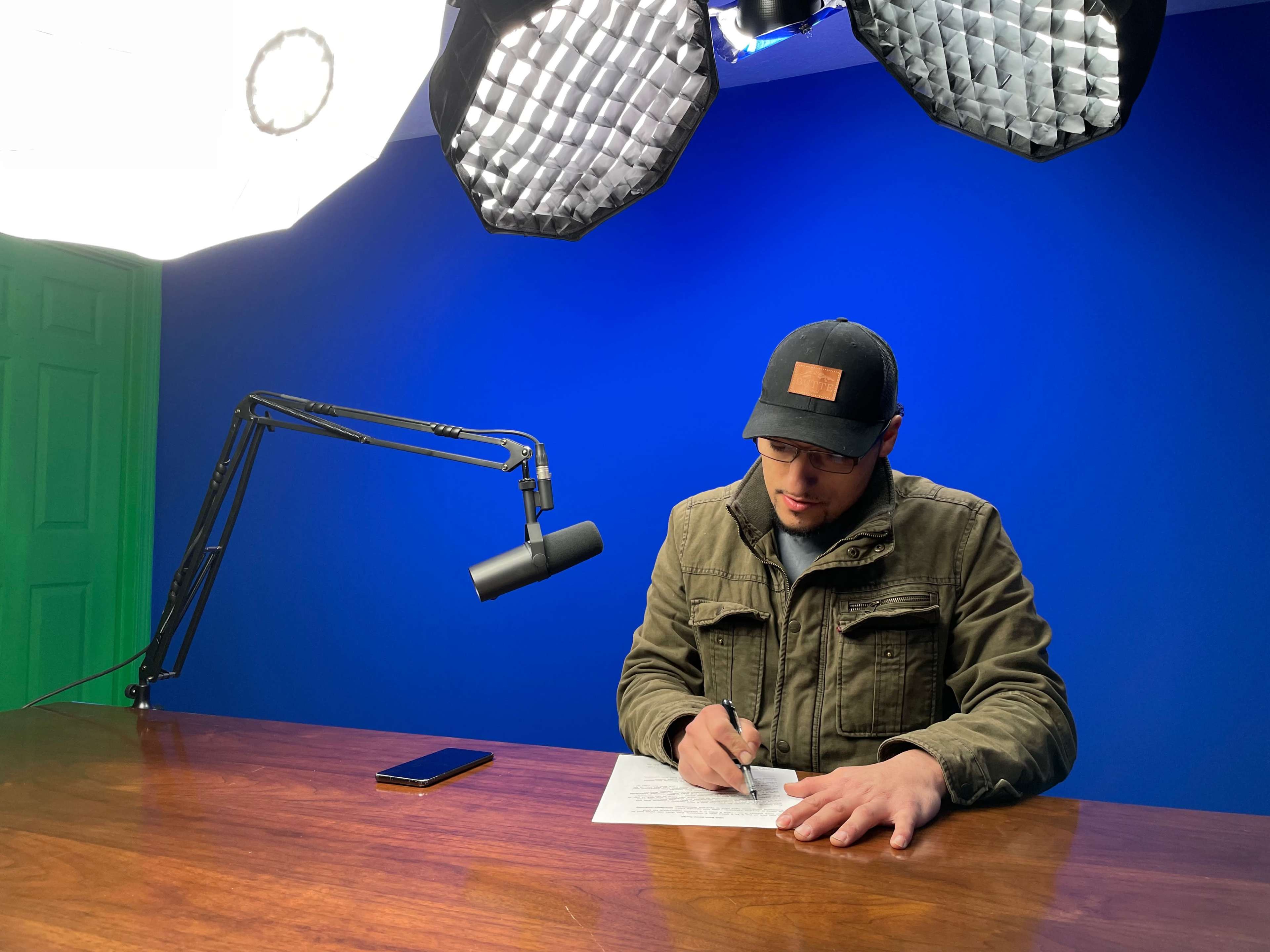 A person wearing a cap writes on paper at a wooden table under bright studio lights, with a blue backdrop and a microphone positioned nearby.