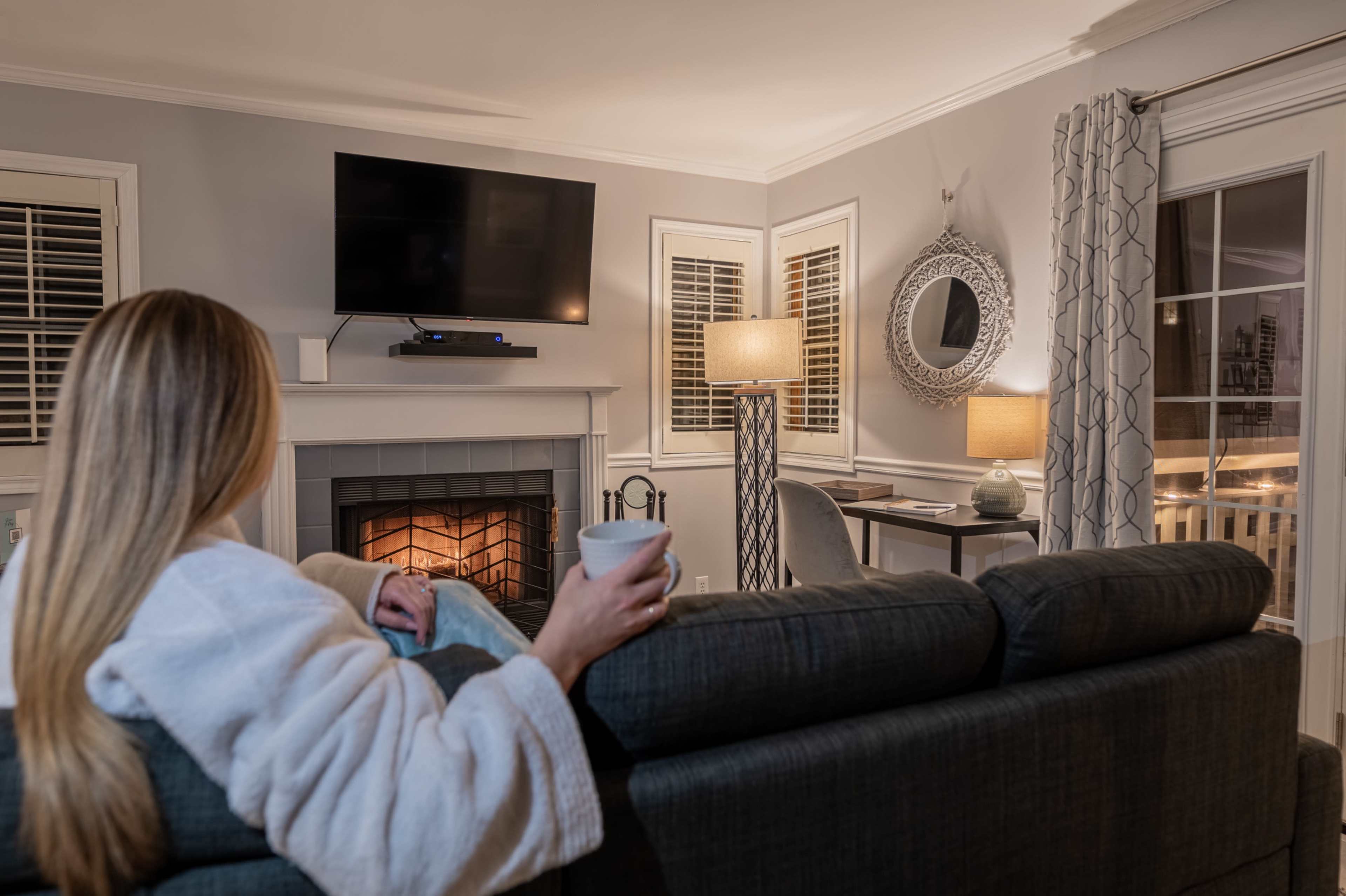 A person in a bathrobe sits on a couch, holding a cup and looking towards a television and a fireplace in a cozy living room.