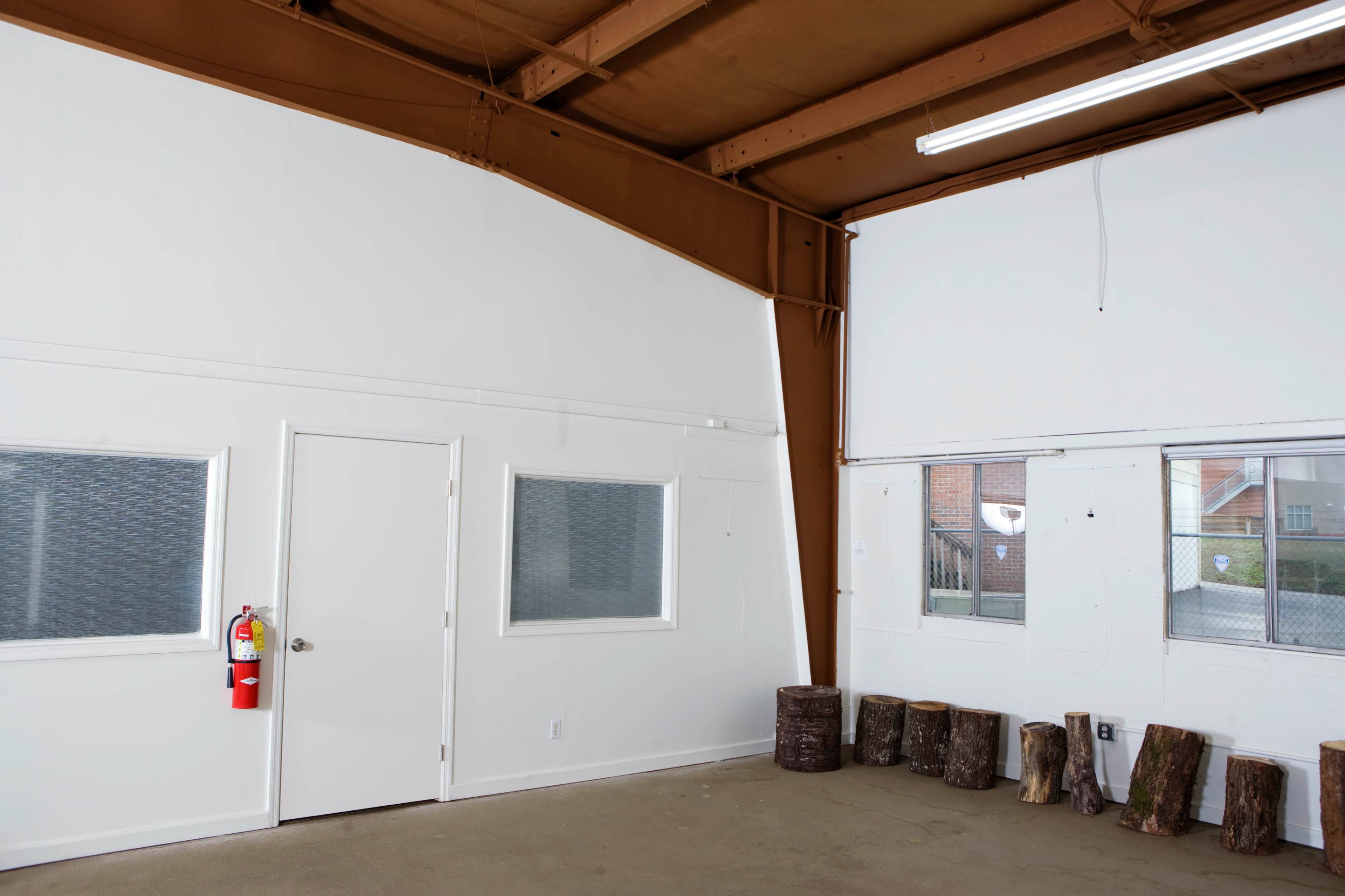The image shows an empty interior space with a white wall, a door, a few windows, and stacked logs along the floor.
