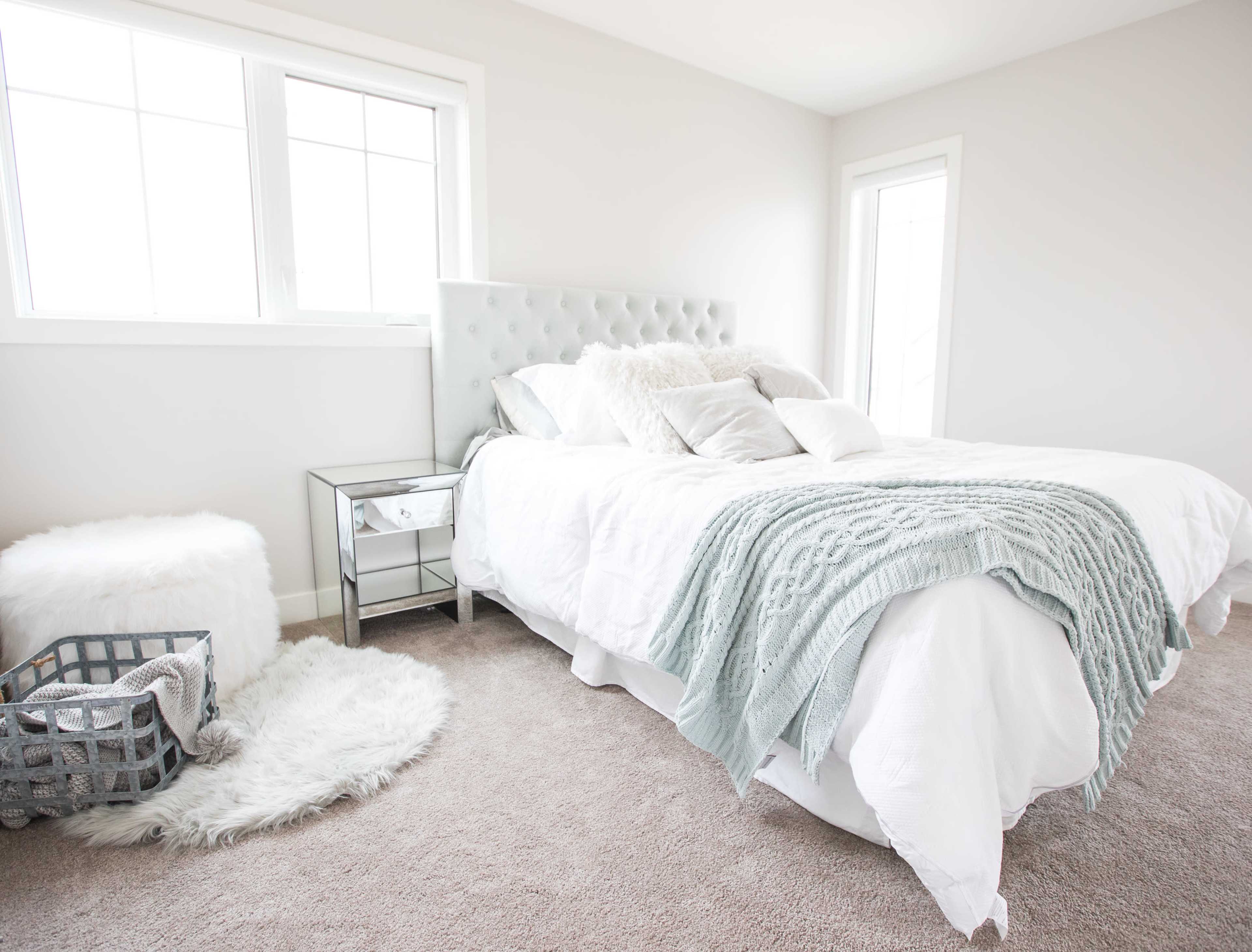 A bright bedroom features a light gray tufted headboard, a white bed with decorative pillows, and a cozy area with a textured throw and a small side table.