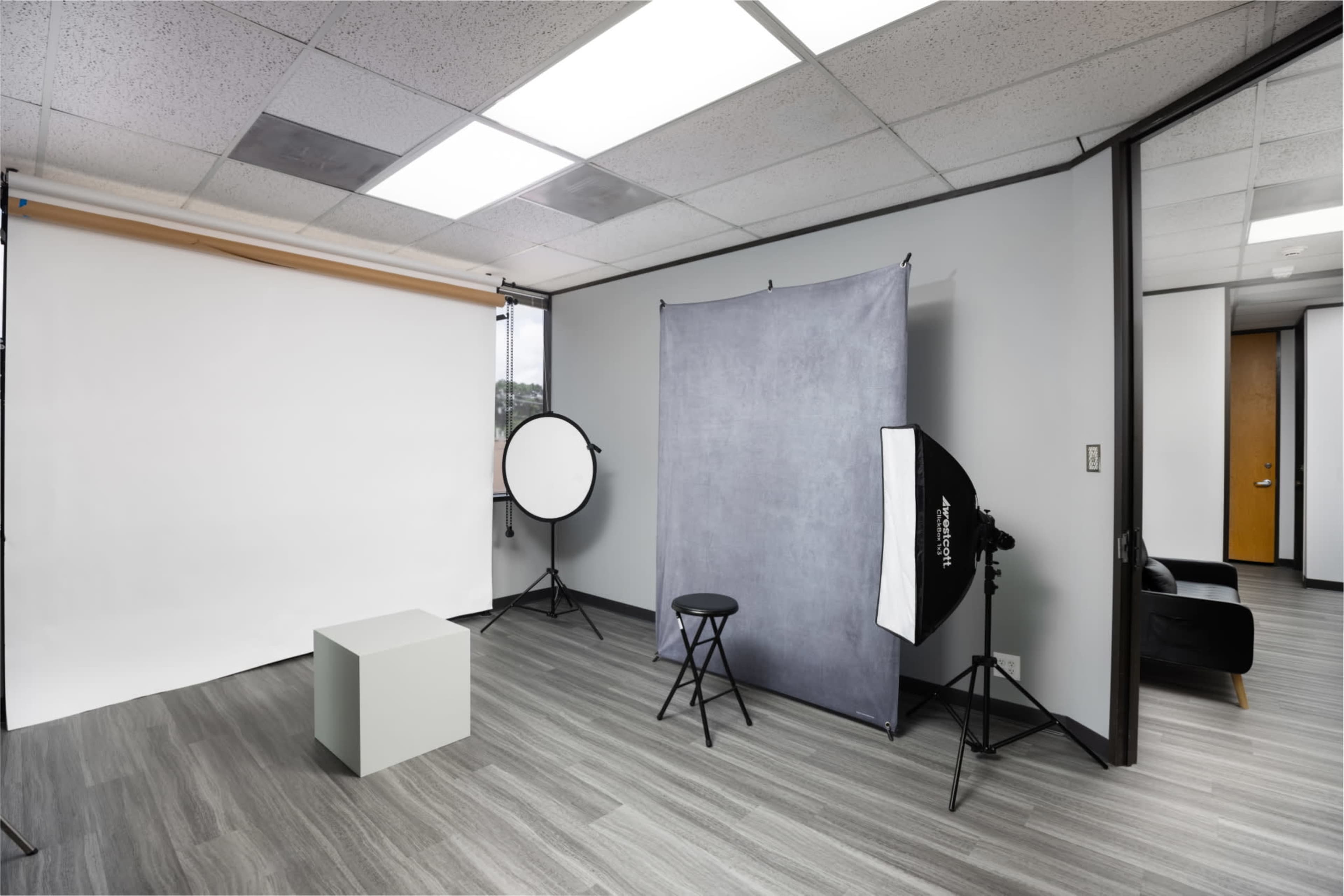 The image shows a photo studio setup featuring a backdrop, lighting equipment, a stool, and a small white cube on a gray floor.