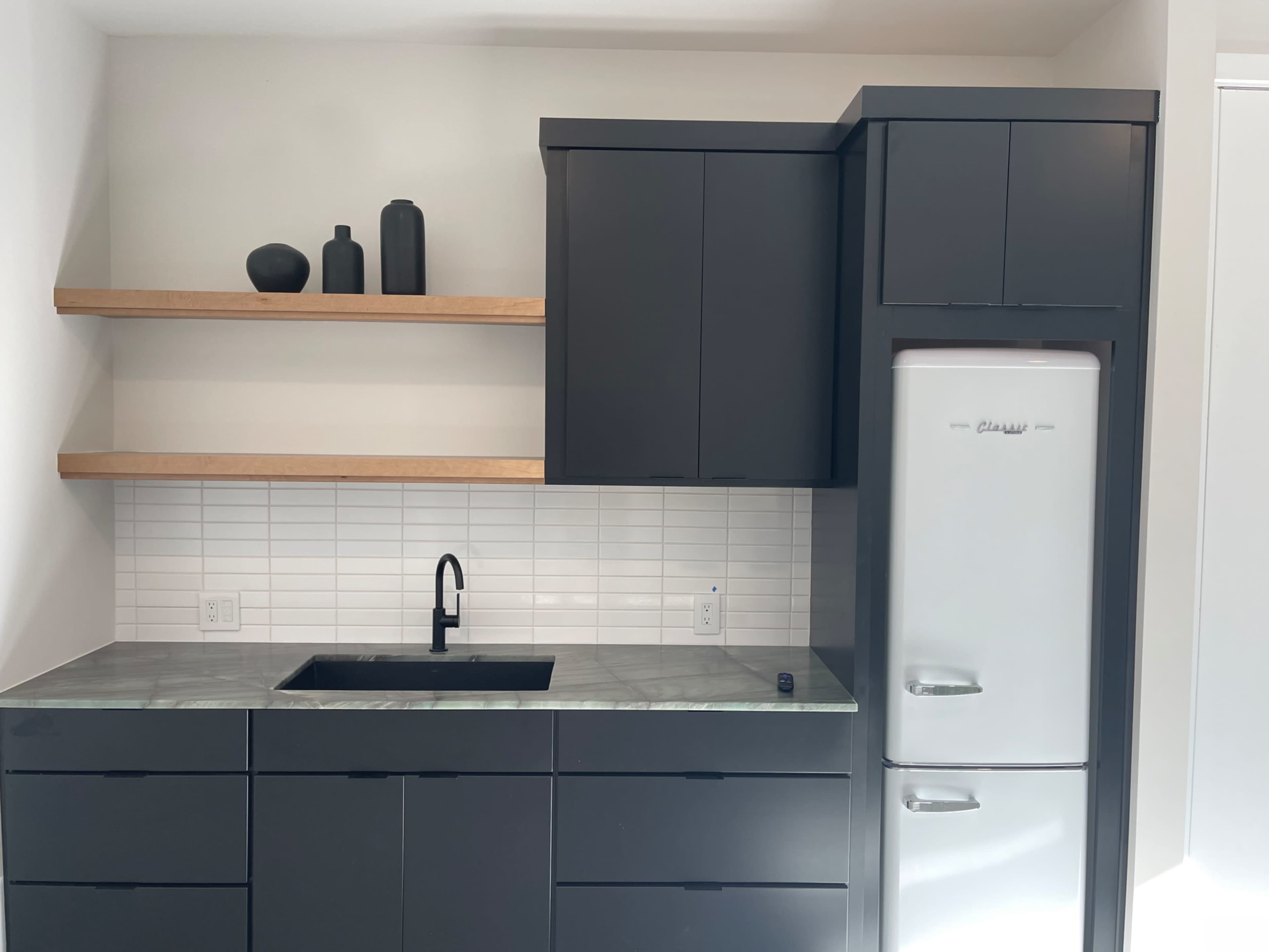 The image shows a modern kitchen with dark cabinetry, a minimalist sink, open wooden shelves holding decorative items, and a silver refrigerator.