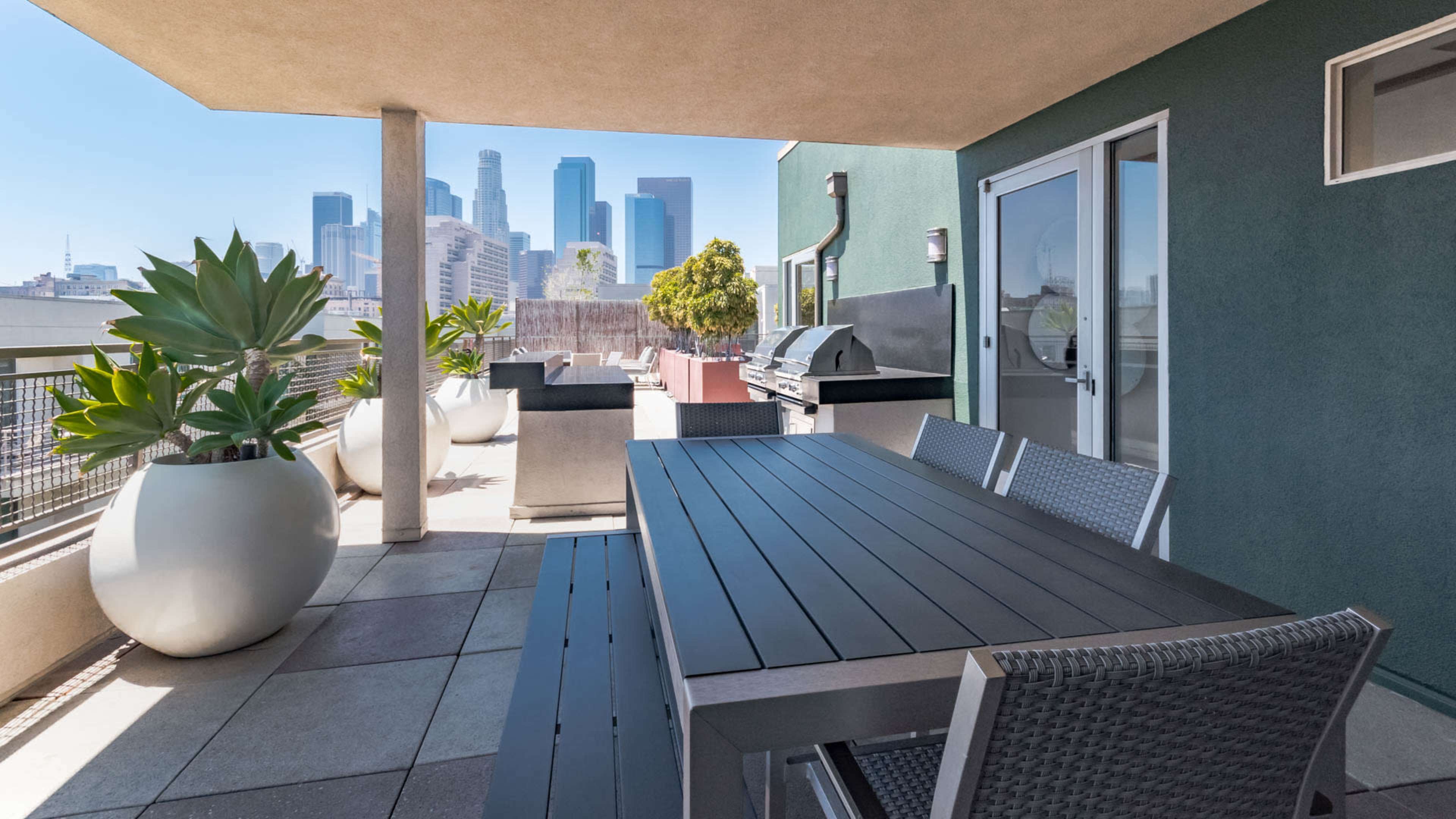 The image shows a spacious balcony with a dining table, grill, and large planters, overlooking a city skyline.