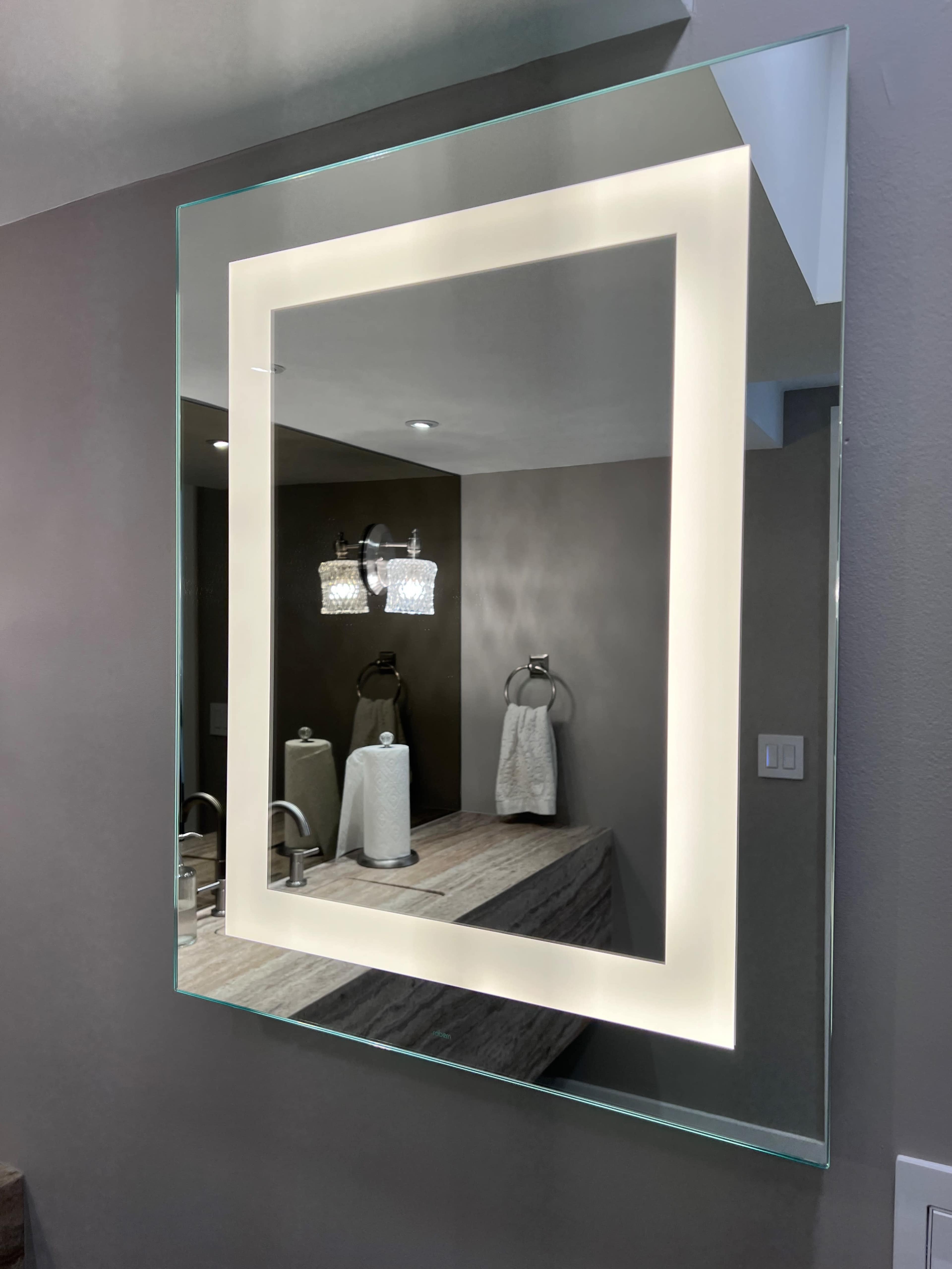 A backlit rectangular mirror mounted on a wall above a countertop with two sinks and towel holders.