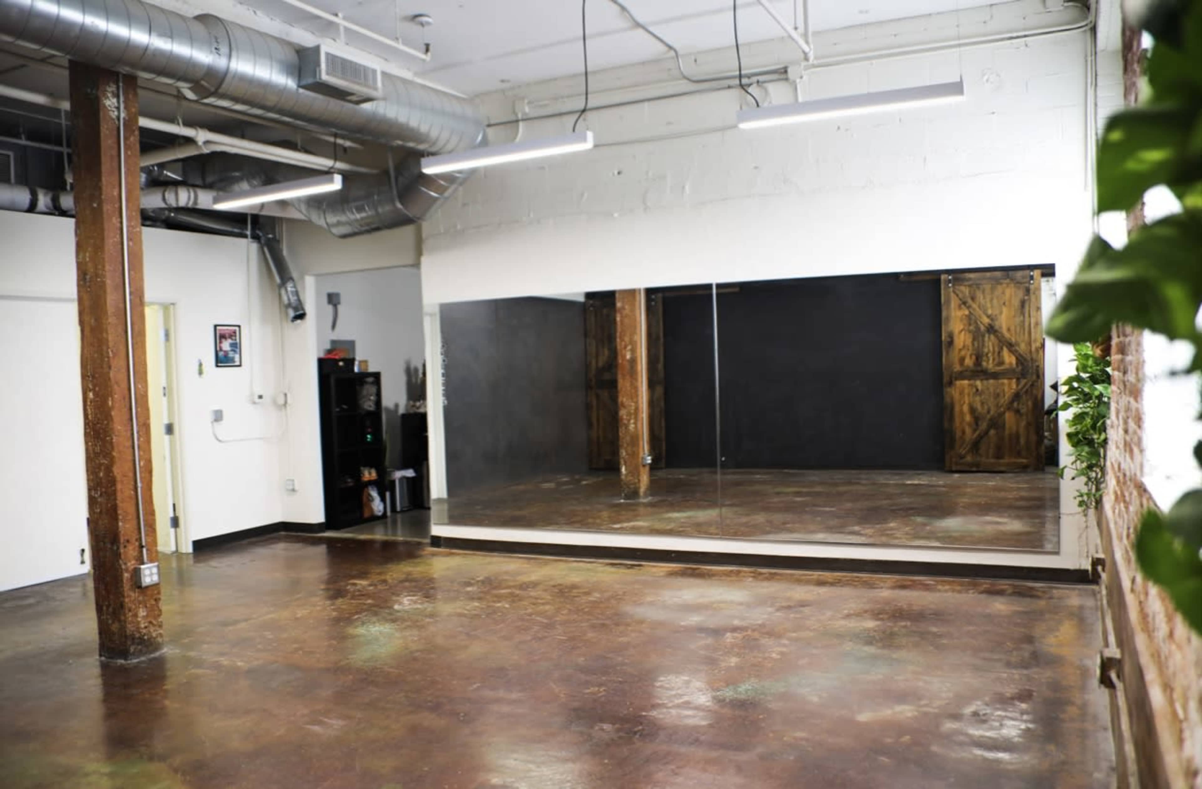 An empty studio space with polished concrete floors, exposed wooden beams, a large mirror on one wall, and a dark accent wall.