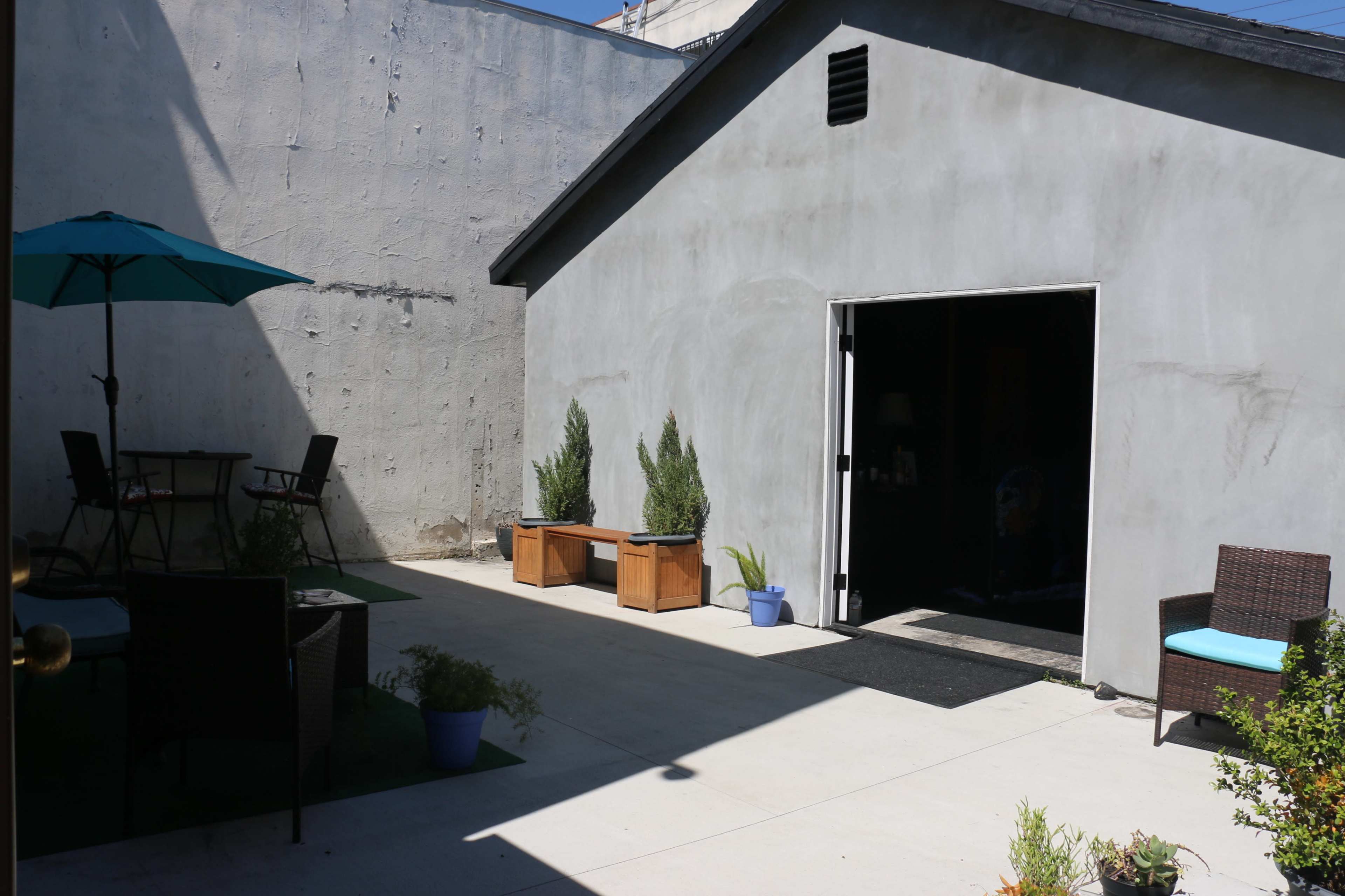 A small outdoor courtyard features a gray wall, a shed, and arranged seating with potted plants.