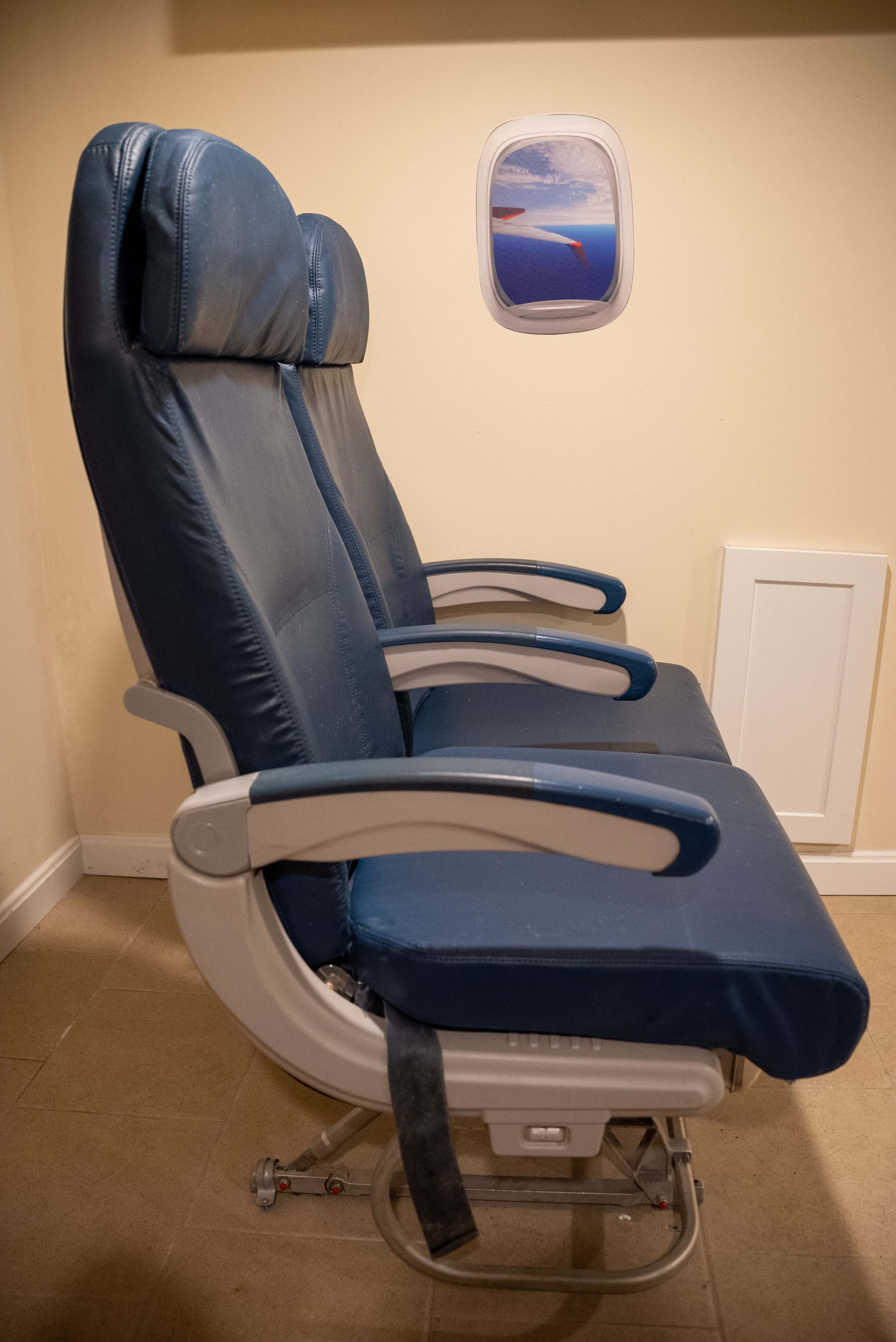 Two blue airplane seats are positioned side by side, accompanied by a window display showing a sky view.