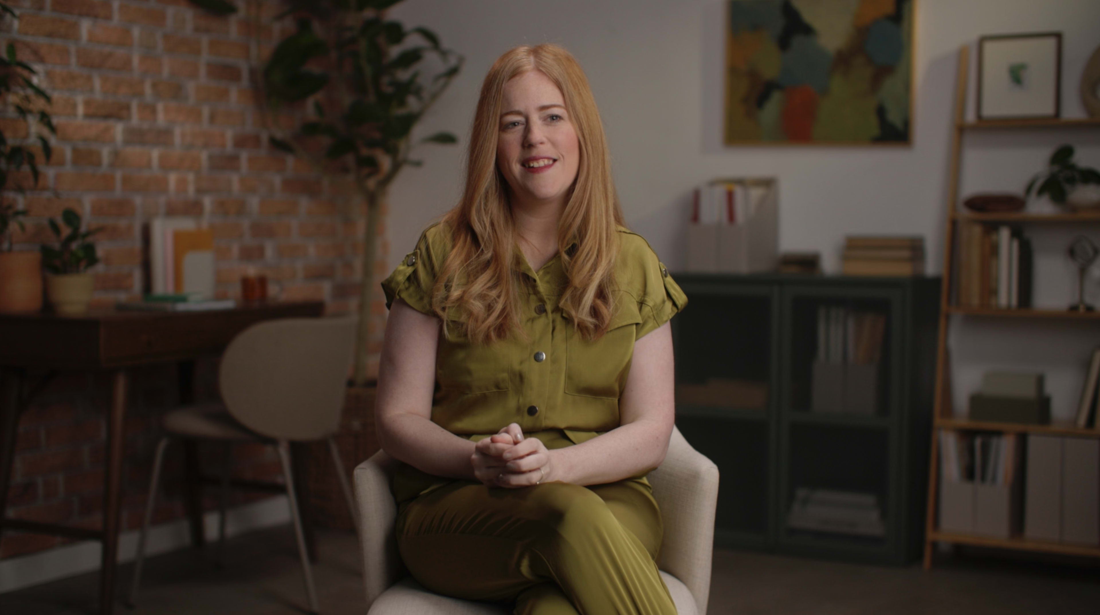 A woman with long red hair sits on a chair in a well-lit room with brick walls and plants, wearing a matching olive-green outfit.