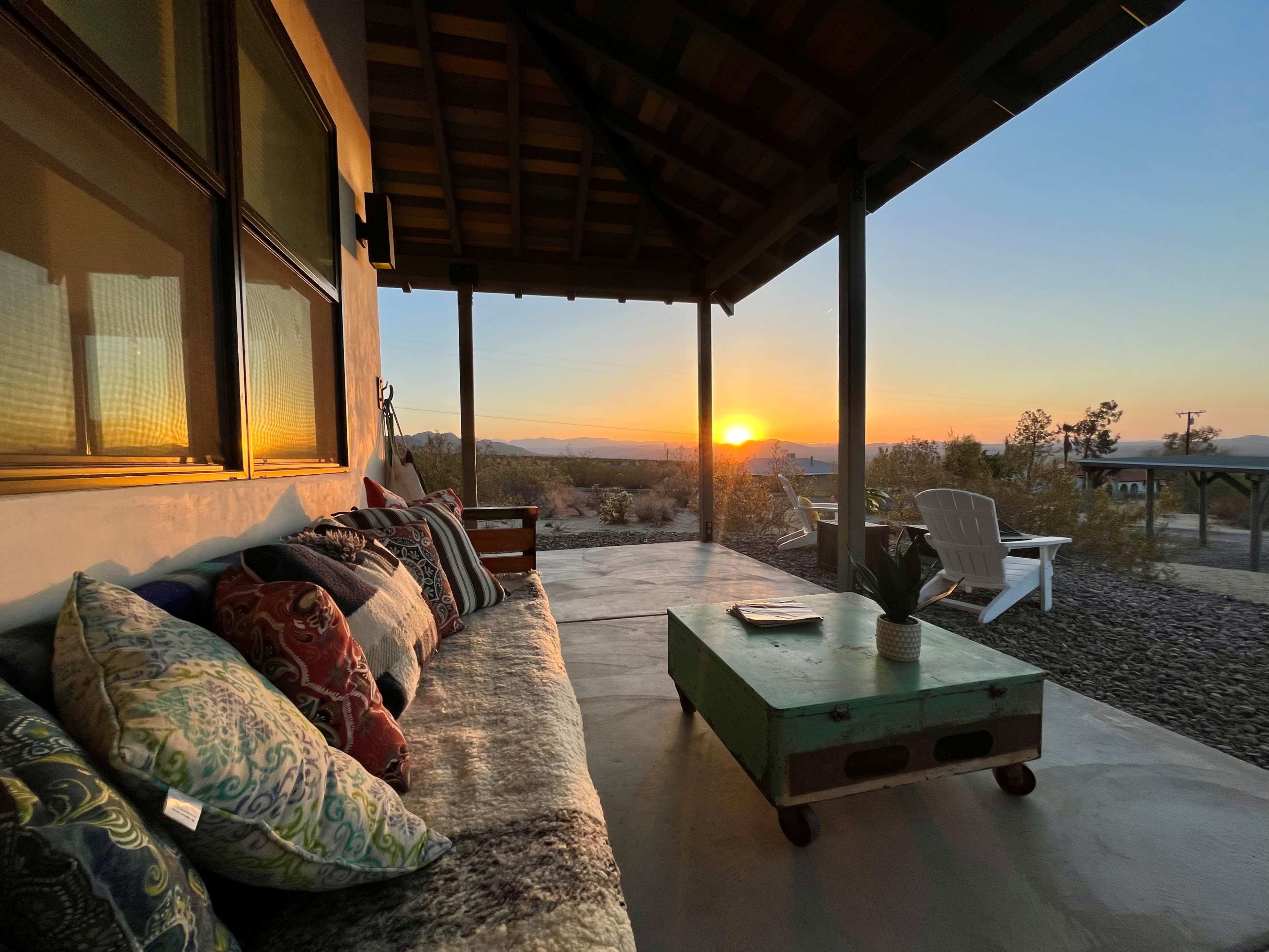 A cozy porch with a colorful couch overlooks a sunset in a desert landscape.