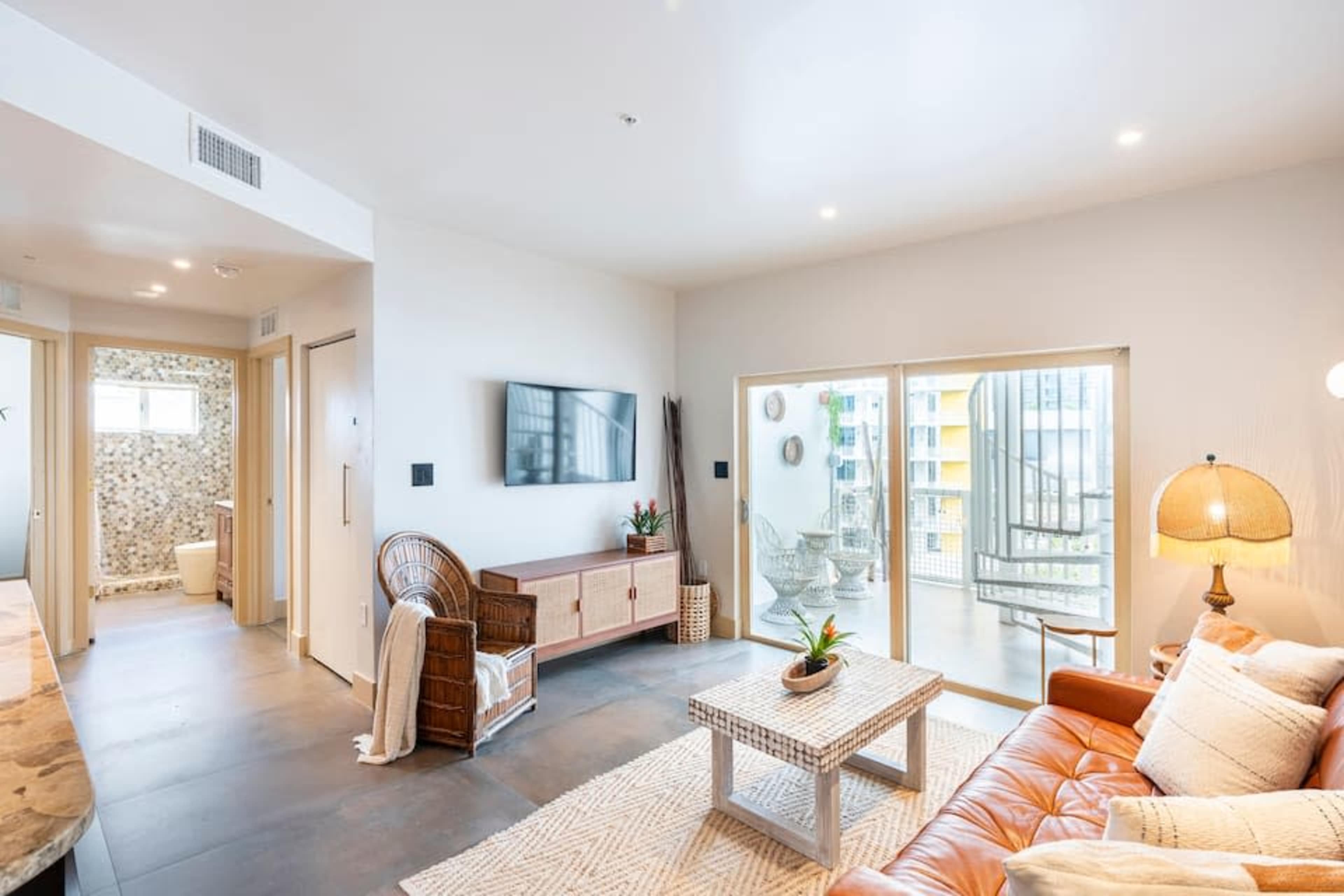 The image shows a modern living room with a brown leather sofa, a coffee table, and large sliding glass doors that open to a balcony, with a light-colored wall and decorative elements.