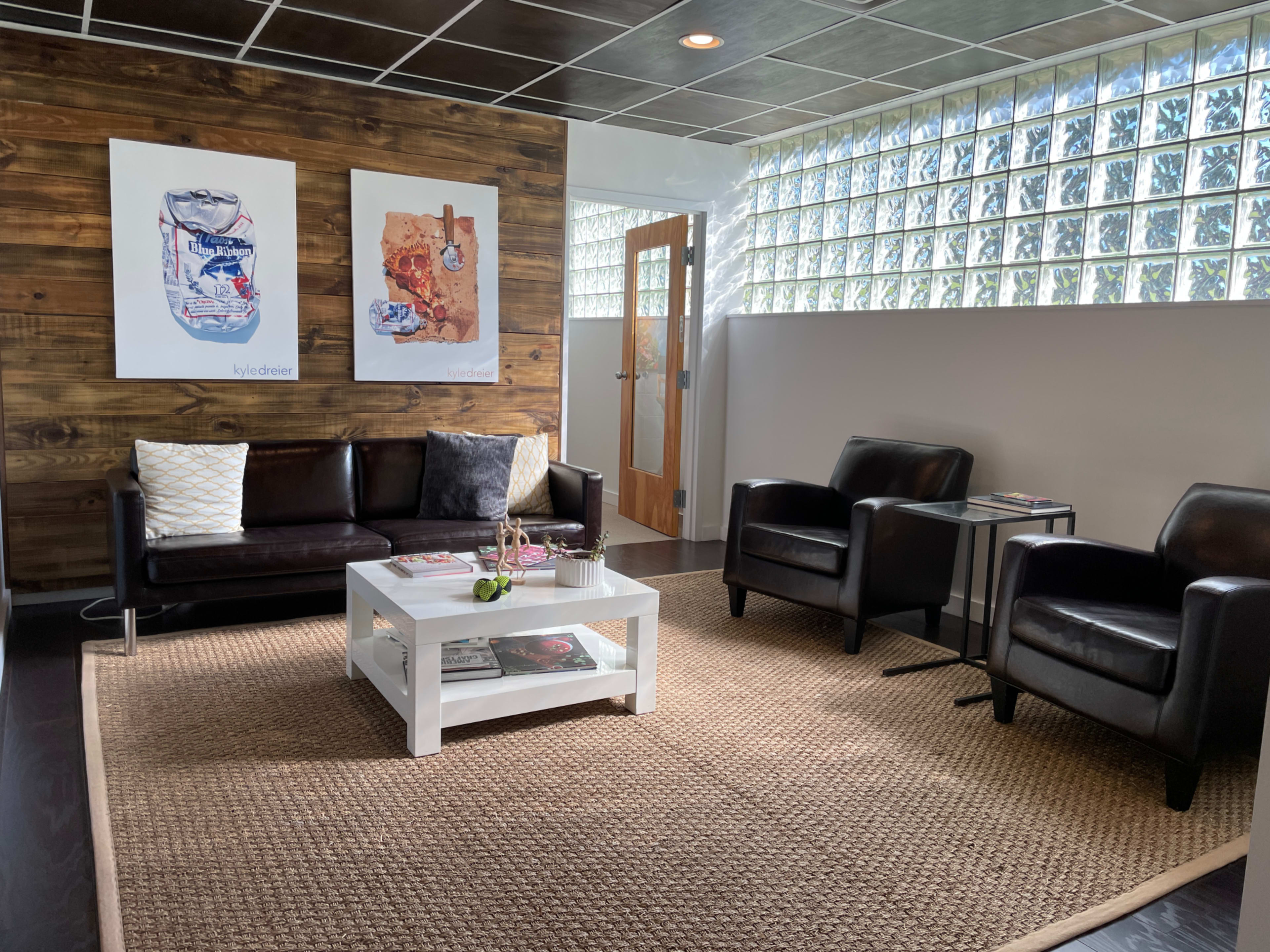 The image shows a modern sitting area with a wooden accent wall, two black chairs, a black couch, and a coffee table on a woven rug.