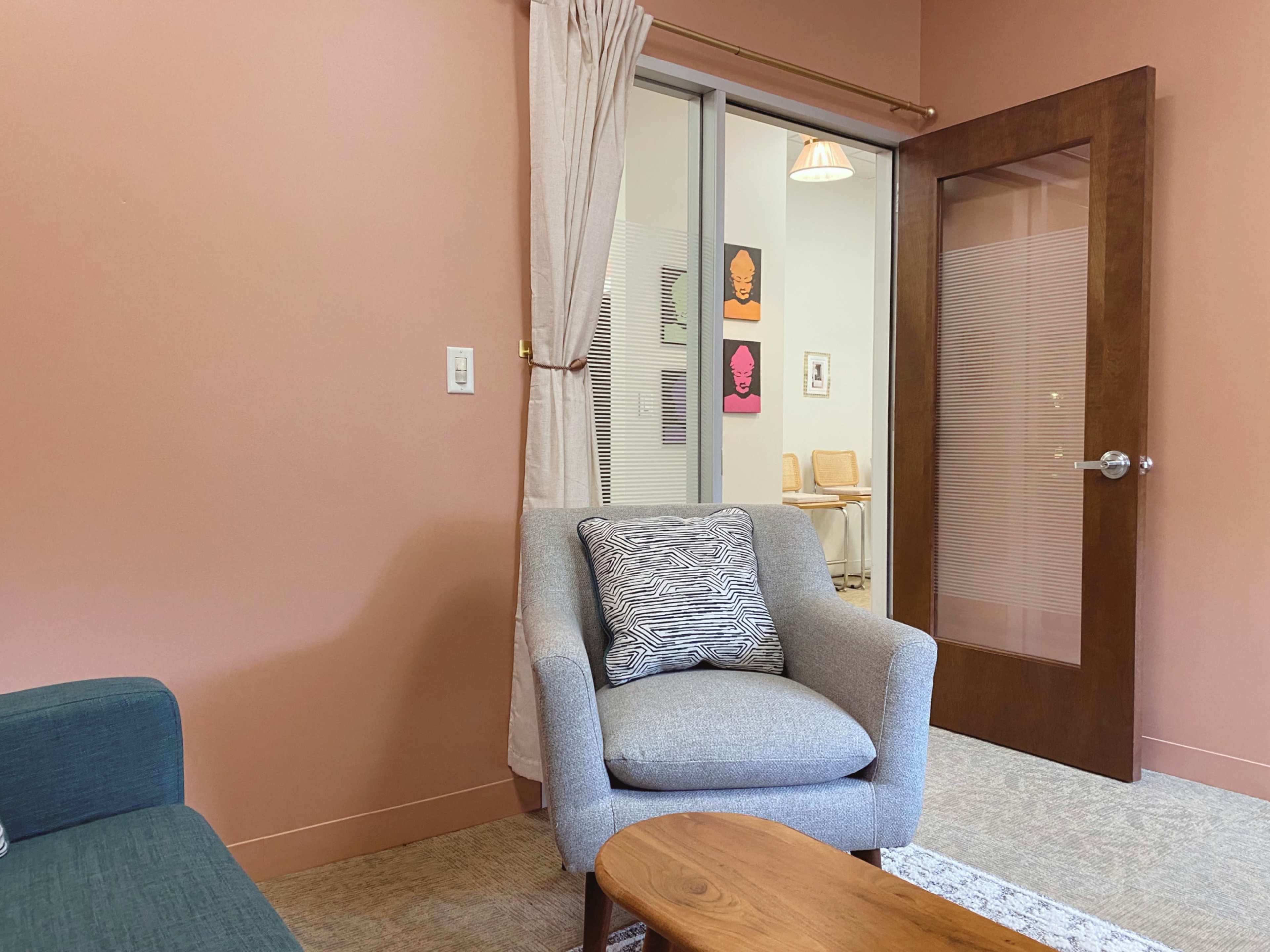 The image shows a cozy indoor space with a gray armchair, a wooden coffee table, and a doorway leading to a well-lit room featuring artwork on the walls.