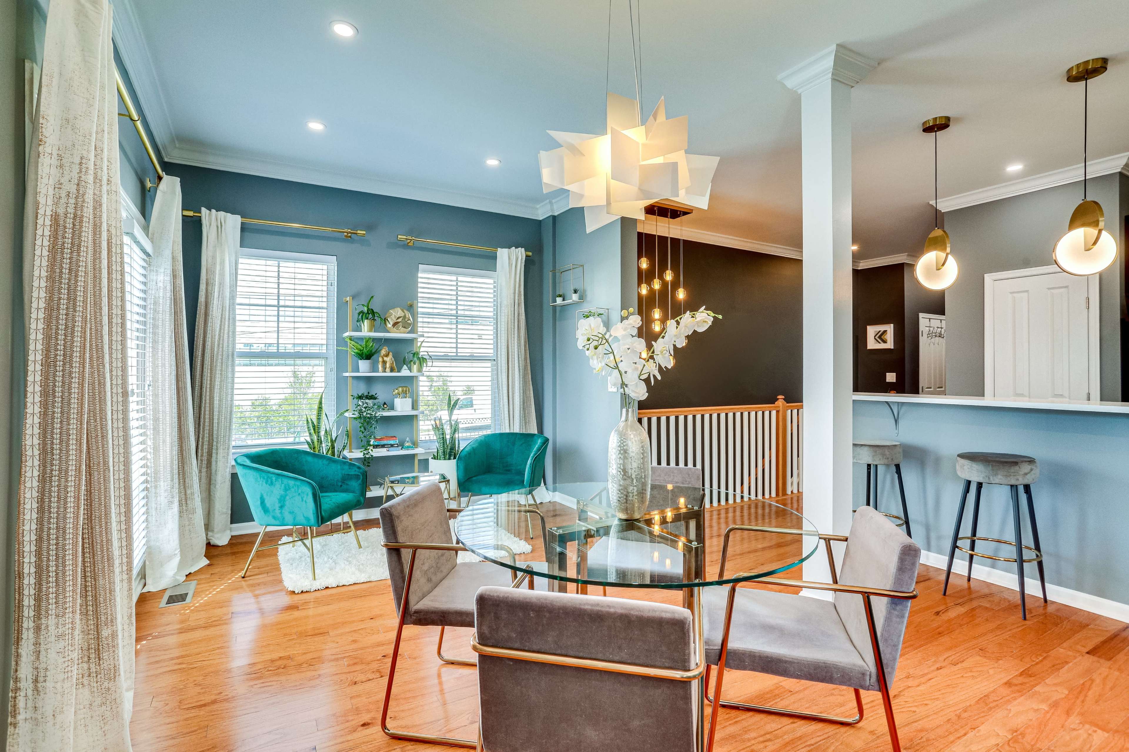 A modern living area with a glass dining table, teal armchairs, and bright natural lighting coming from large windows.