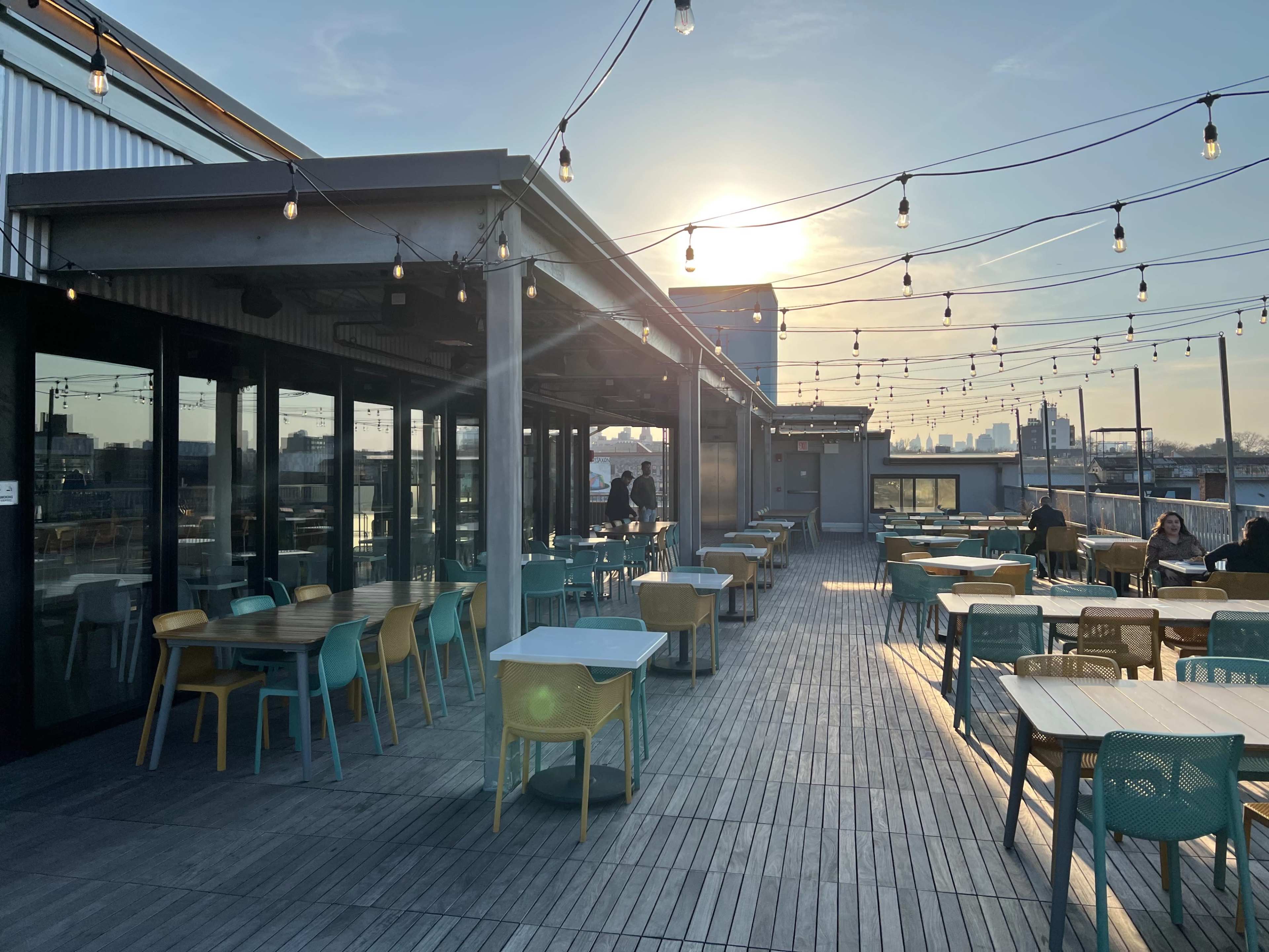 A rooftop space features rows of tables and chairs under string lights, with the sun setting in the background.