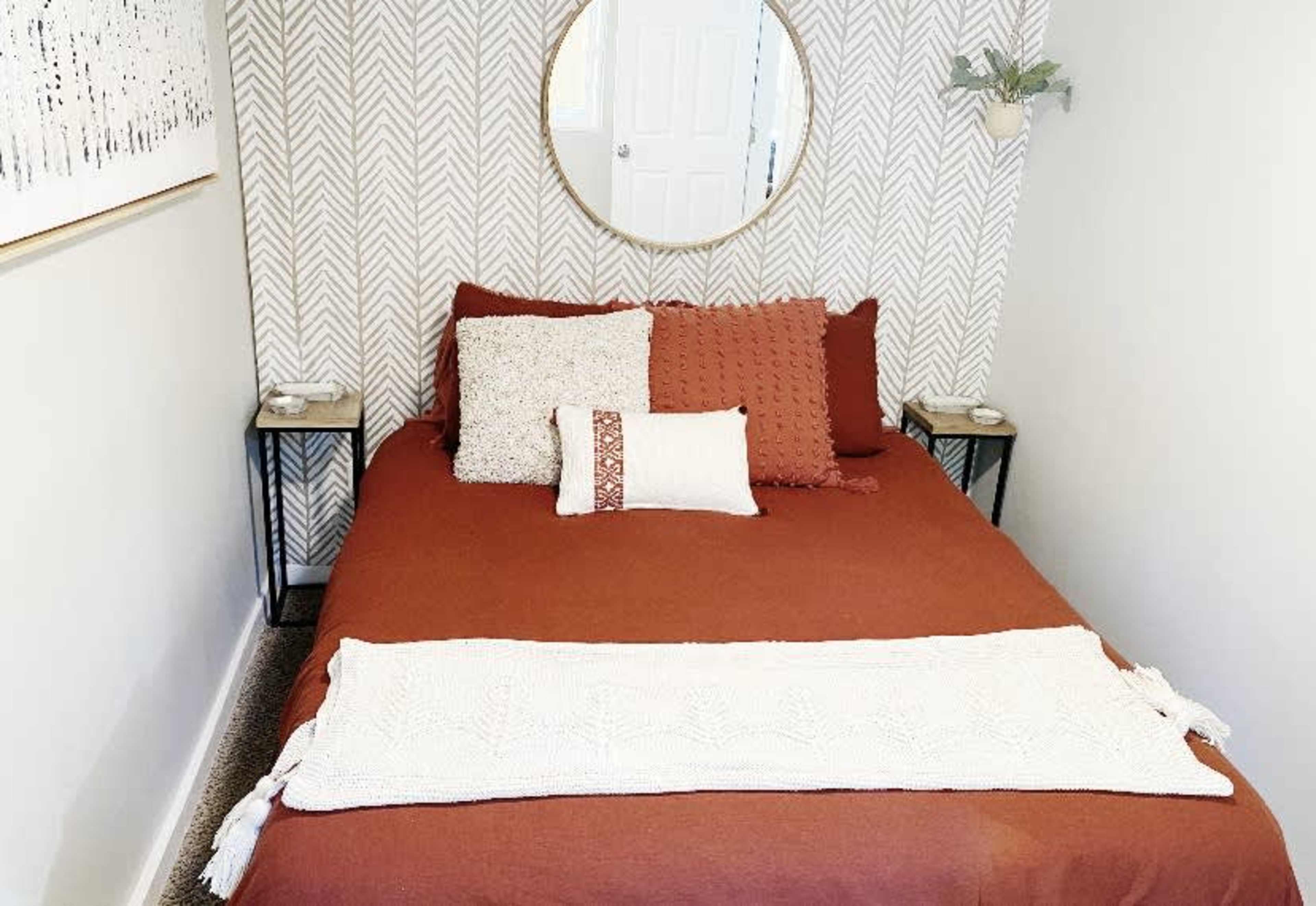 A neatly made bed with a rust-colored duvet and various decorative pillows, positioned against a patterned wall and accompanied by small side tables.