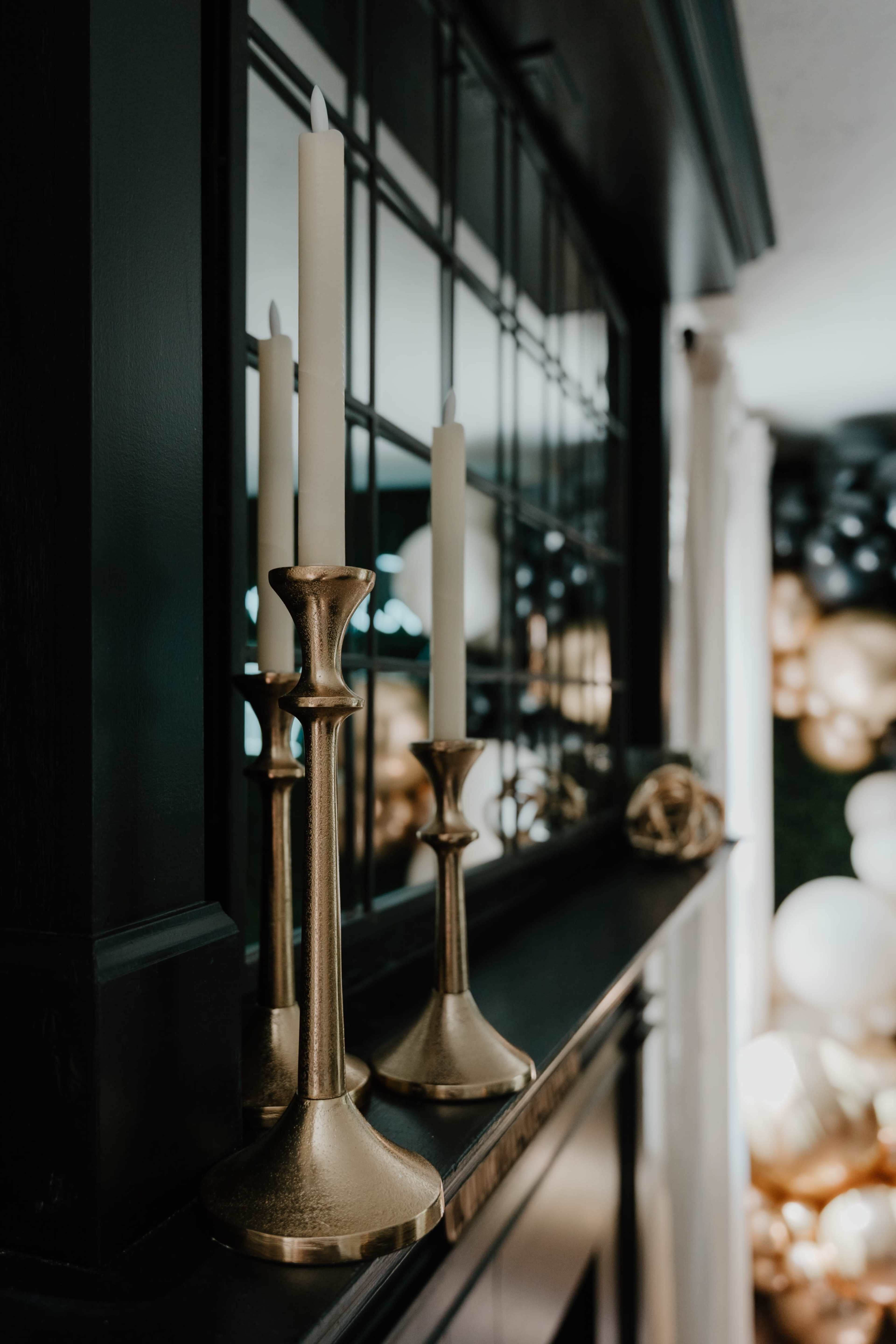 A set of three golden candlesticks with white candles standing on a dark wooden mantel, alongside a backdrop of blurred decorative balloons.