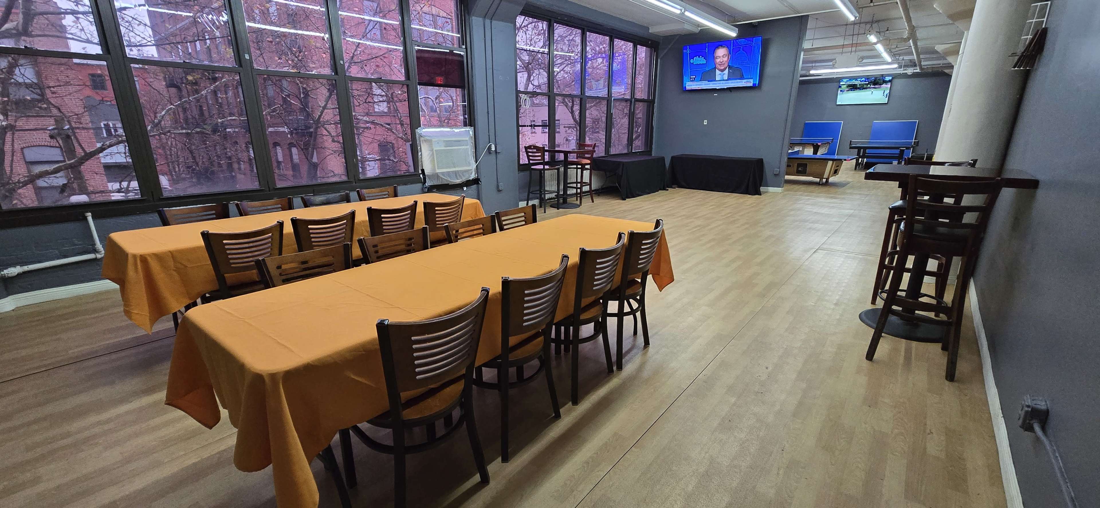 The image shows a spacious interior with several tables and chairs set up, including a long orange table, along with two screens displaying broadcasts.