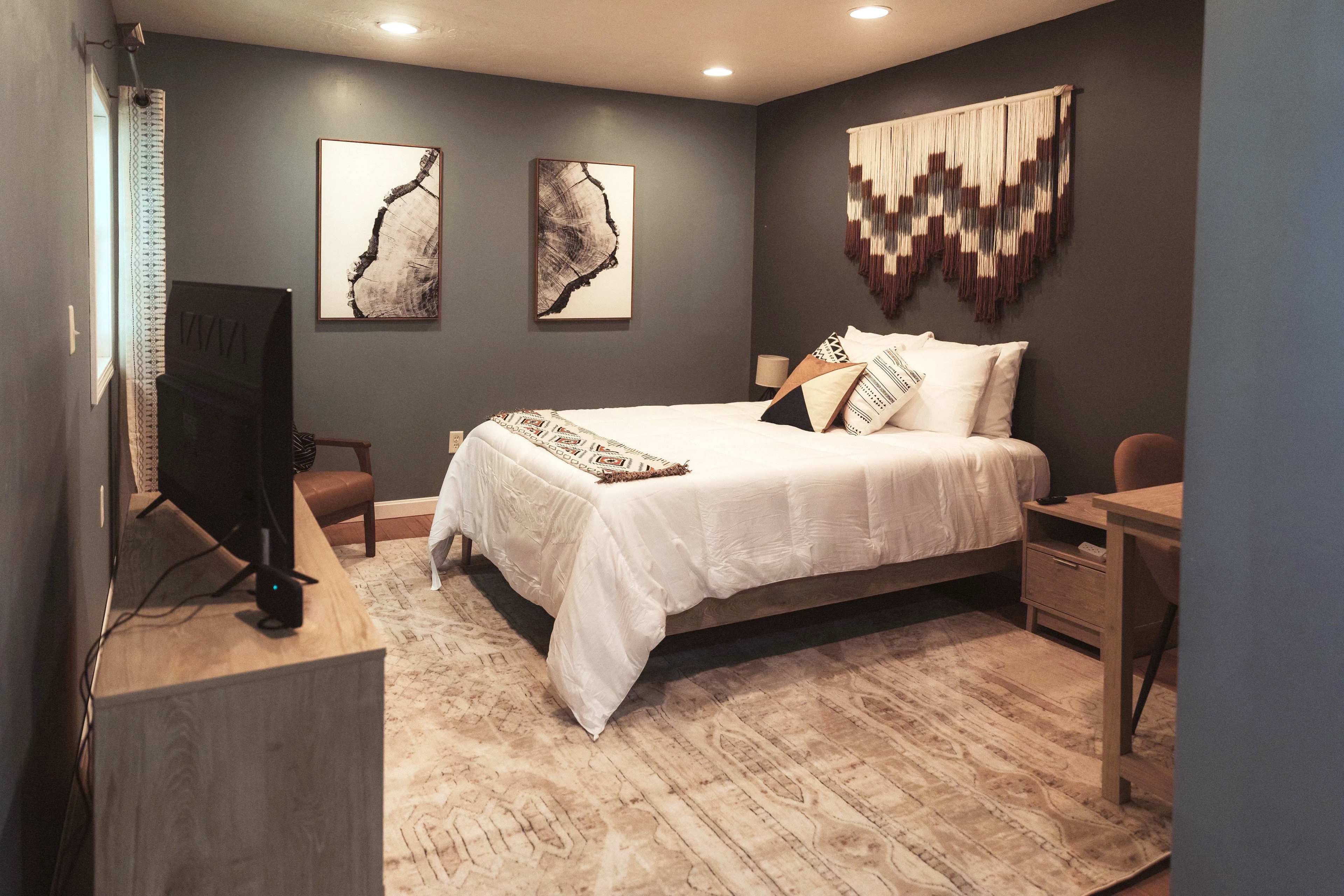 A cozy bedroom featuring a king-sized bed, a bedside table, a wooden dresser with a television, and two abstract wall artworks.
