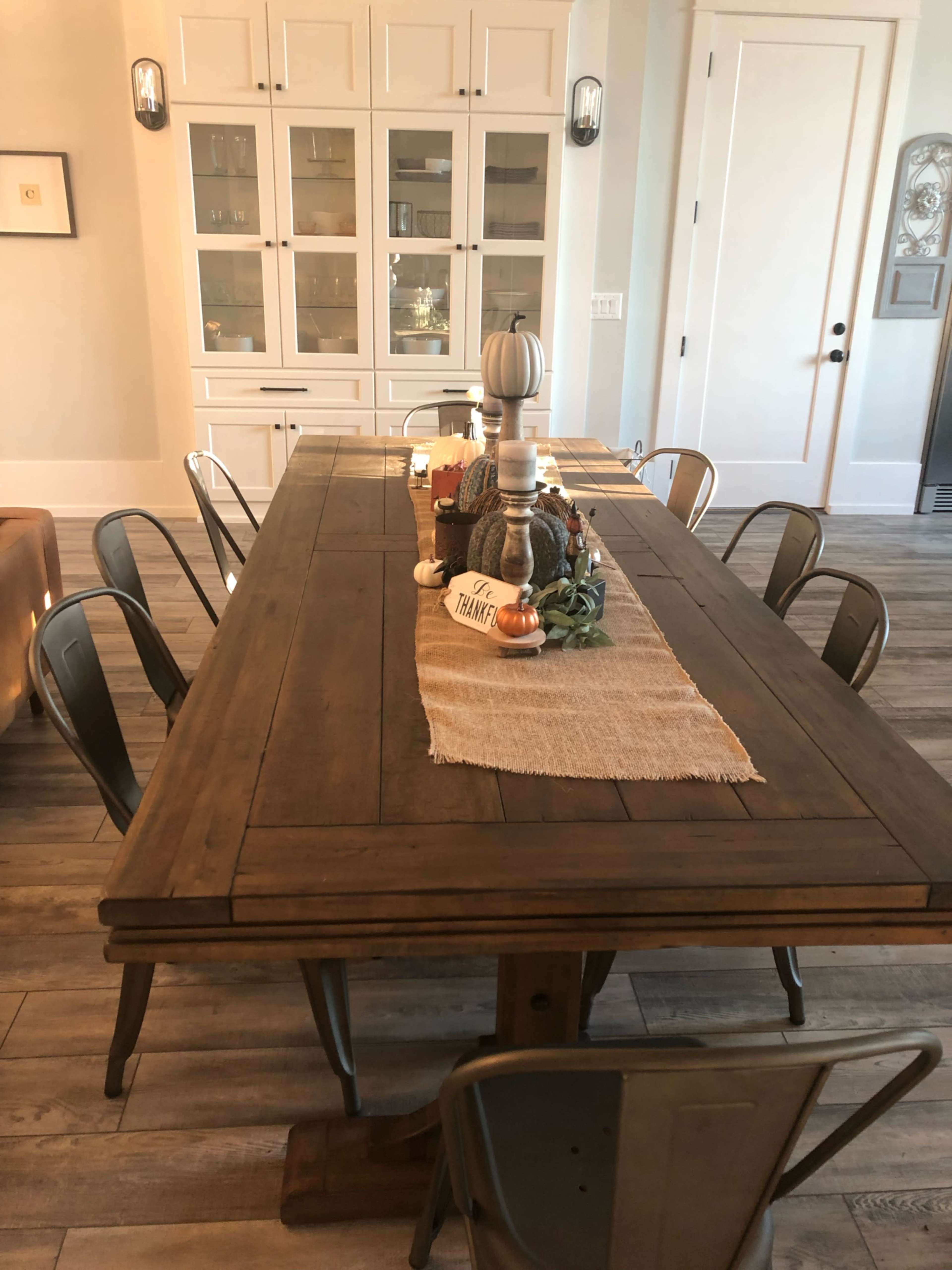 The image shows a wooden dining table set with a center runner and decorative items, surrounded by metal chairs in a bright, spacious room.