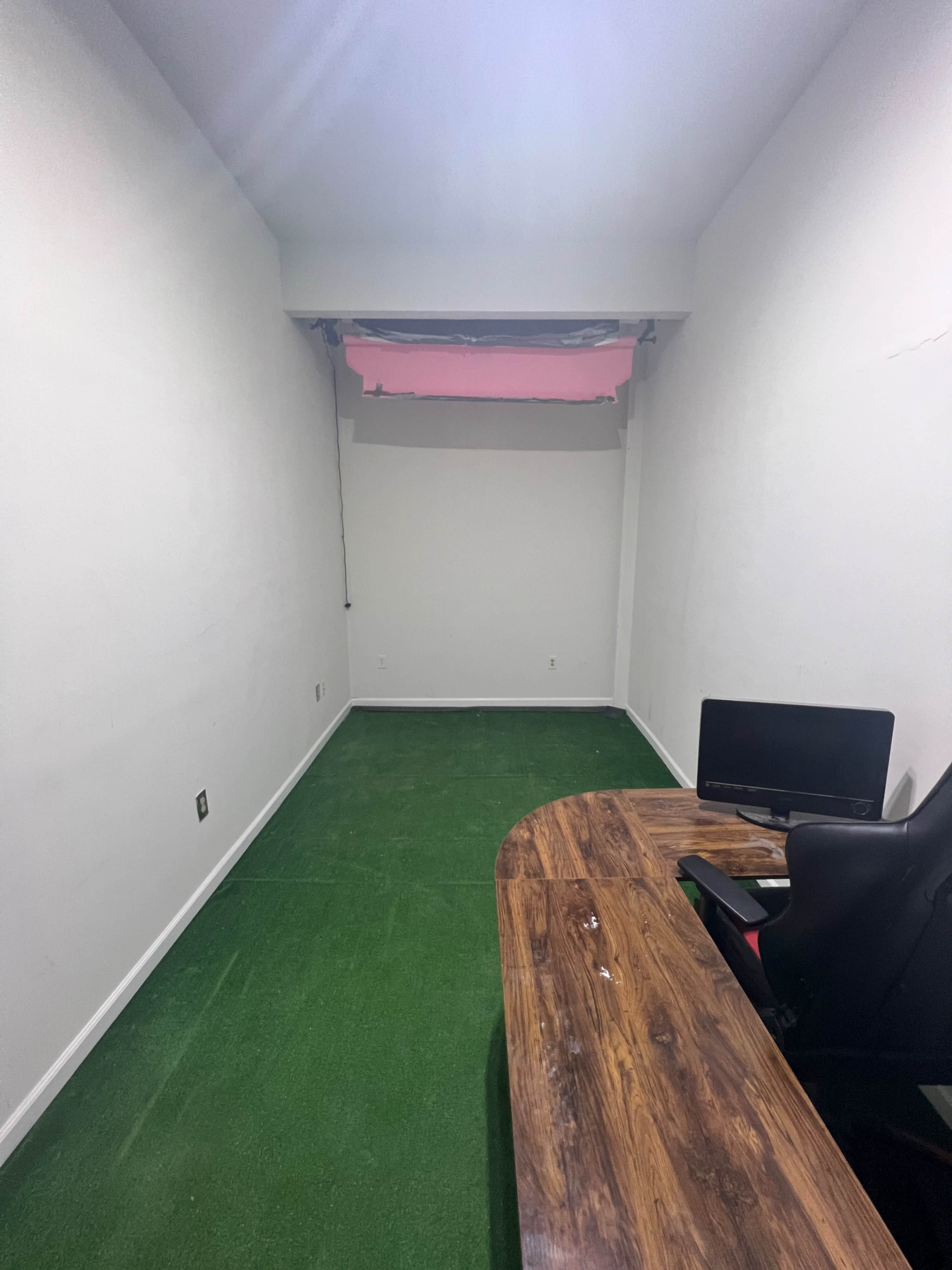 A small, empty room with green carpet, a wooden desk in the corner, a black chair, and a computer monitor.