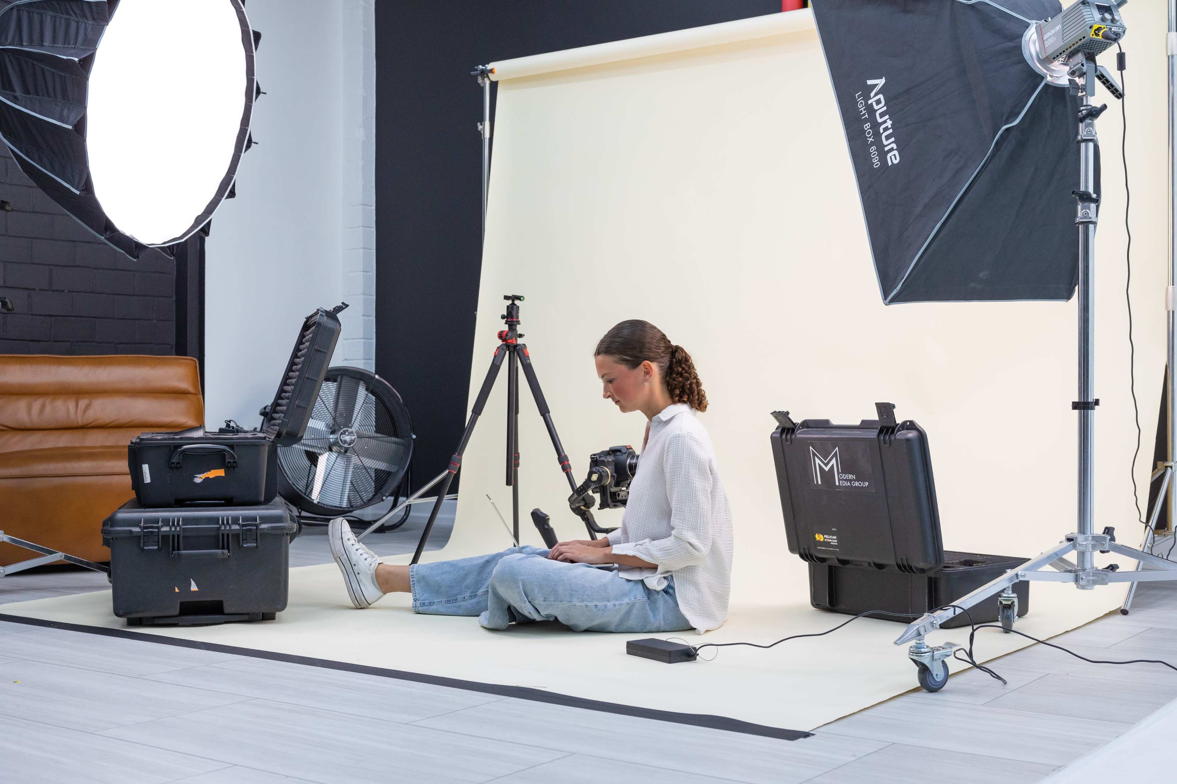 A person sits on a light-colored floor in a photography studio surrounded by camera equipment and lighting gear.