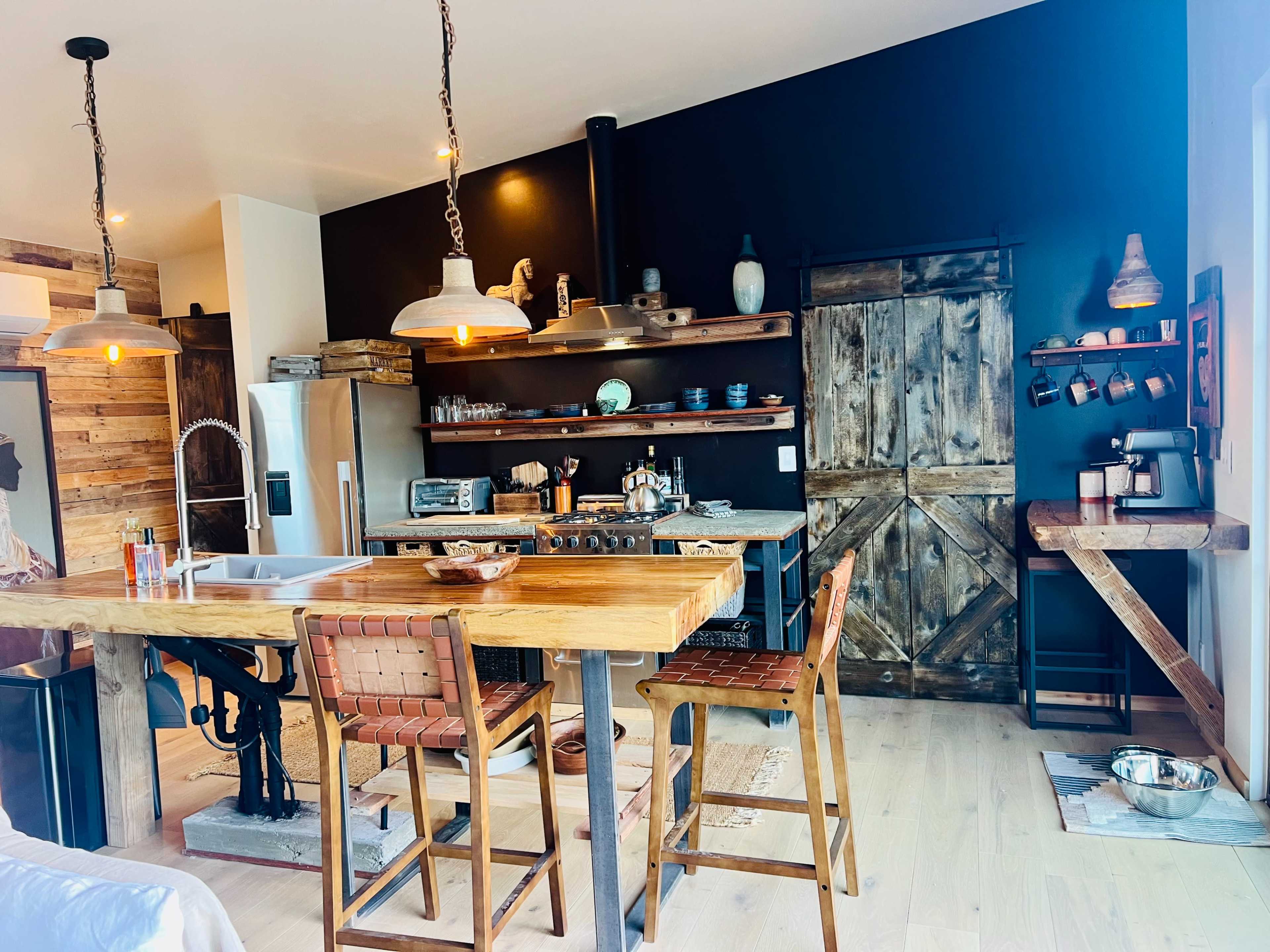 The kitchen features a wooden dining table with metal bar stools, open shelves displaying dishware, and a rustic door leading to another space.