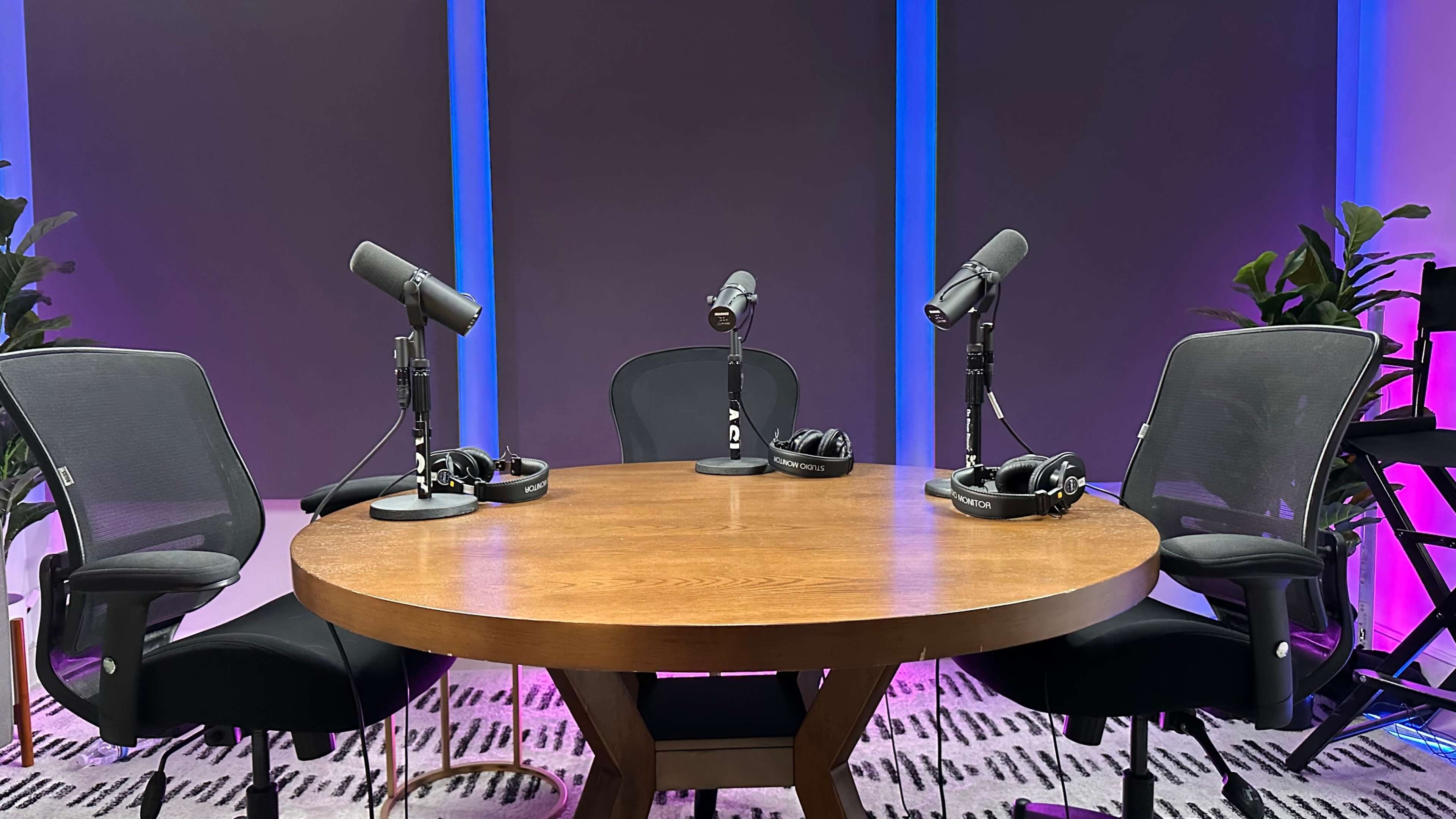 A round wooden table sits in the center of a brightly lit studio, surrounded by four chairs with microphones and headphones set up for a recording session.