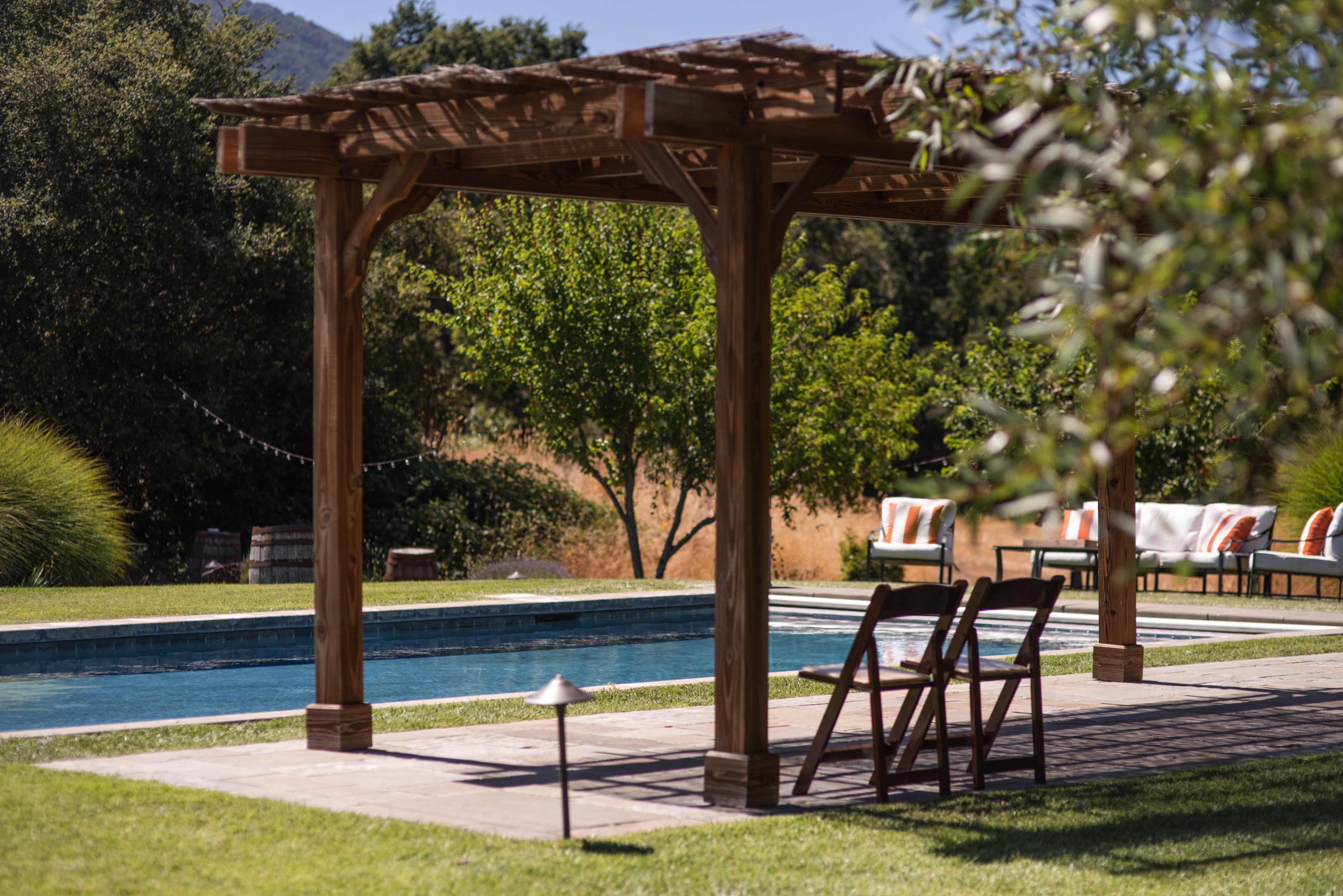 A wooden pergola frames a swimming pool surrounded by landscaped greenery and lounge chairs.