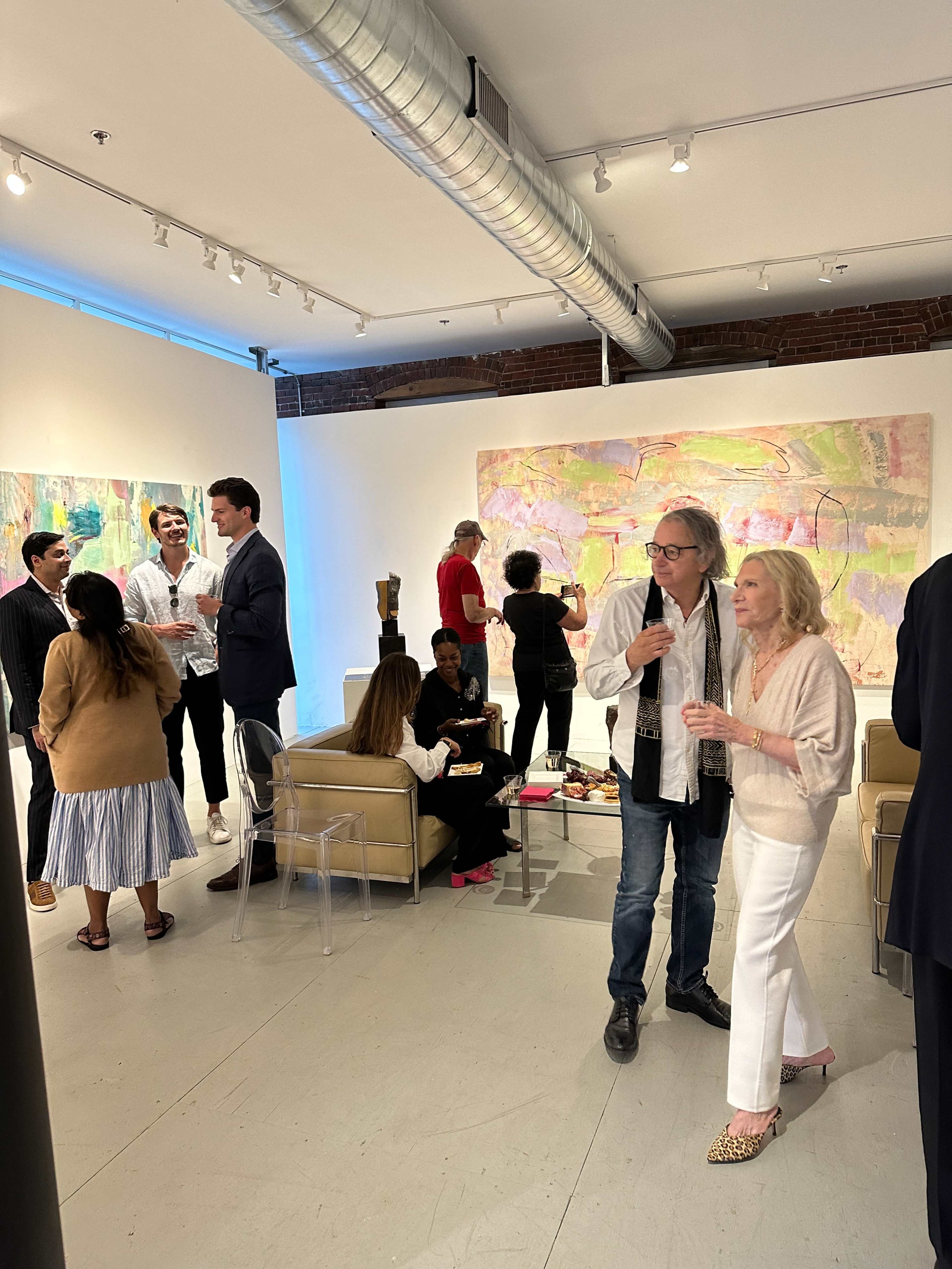 A group of people socialize during an art exhibit in a well-lit gallery featuring colorful abstract paintings on the walls.